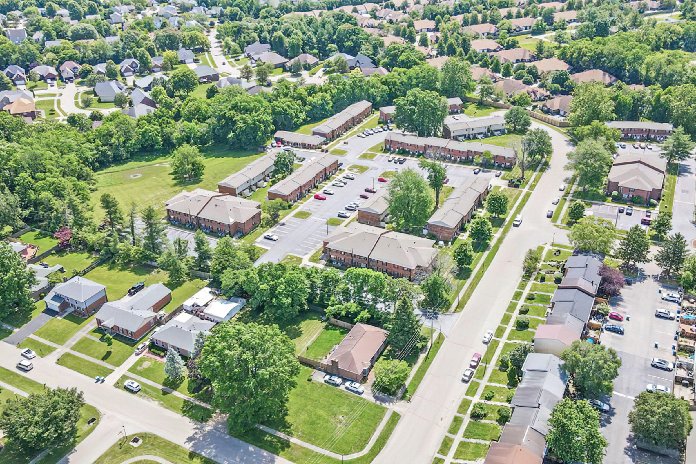 Aerial view of a suburban neighborhood featuring various apartment complexes surrounded by lush greenery. The layout includes parking spaces, trees, and residential streets. The scene captures a mix of buildings, lawns, and driveways, showcasing a typical community setting.