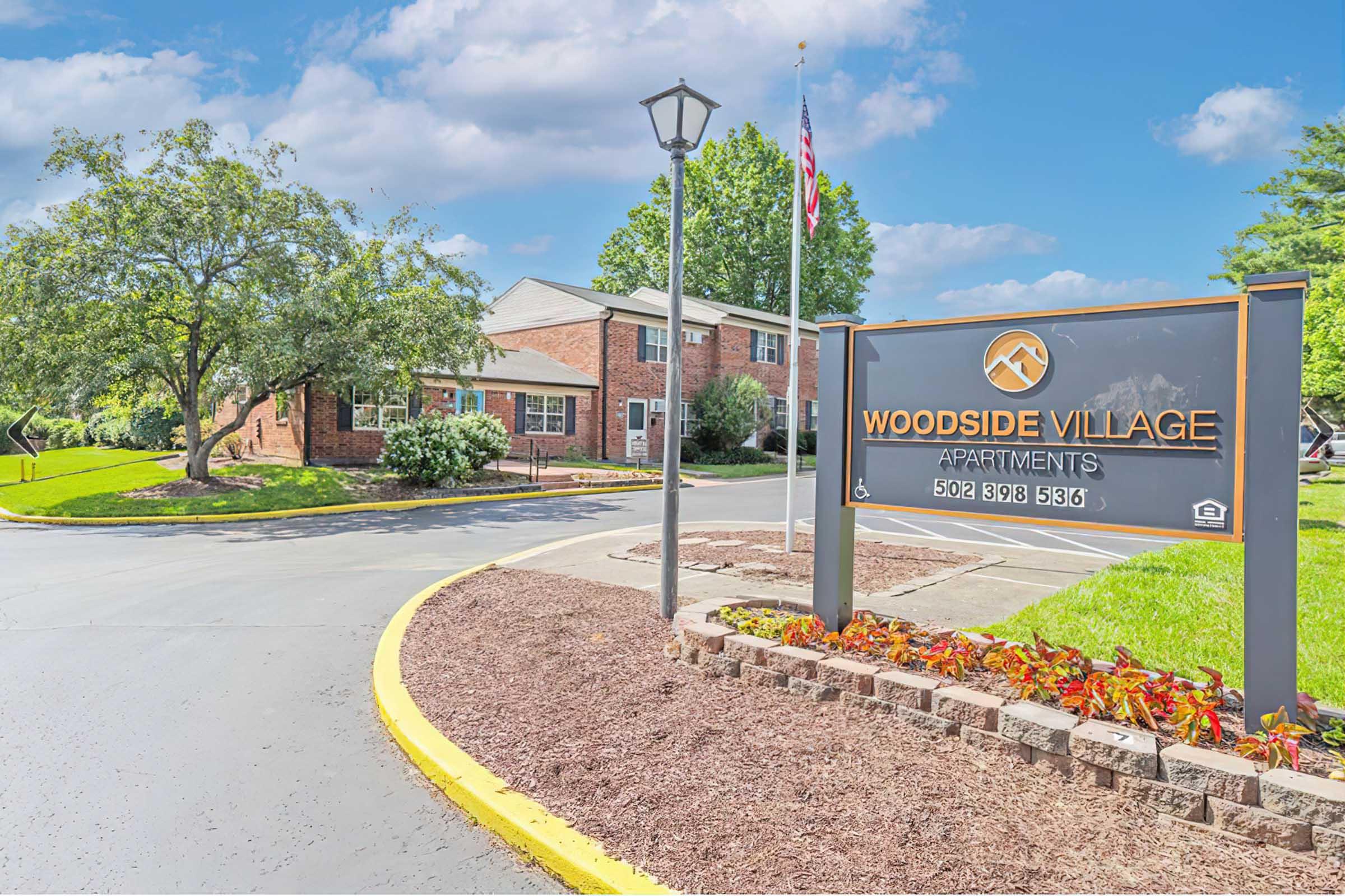 Exterior view of Woodside Village Apartments featuring red brick buildings, well-manicured landscaping, and a sign showing the name of the apartment complex. A lamppost and flagpole are visible, with a curved driveway leading into the property. The sky is partly cloudy.