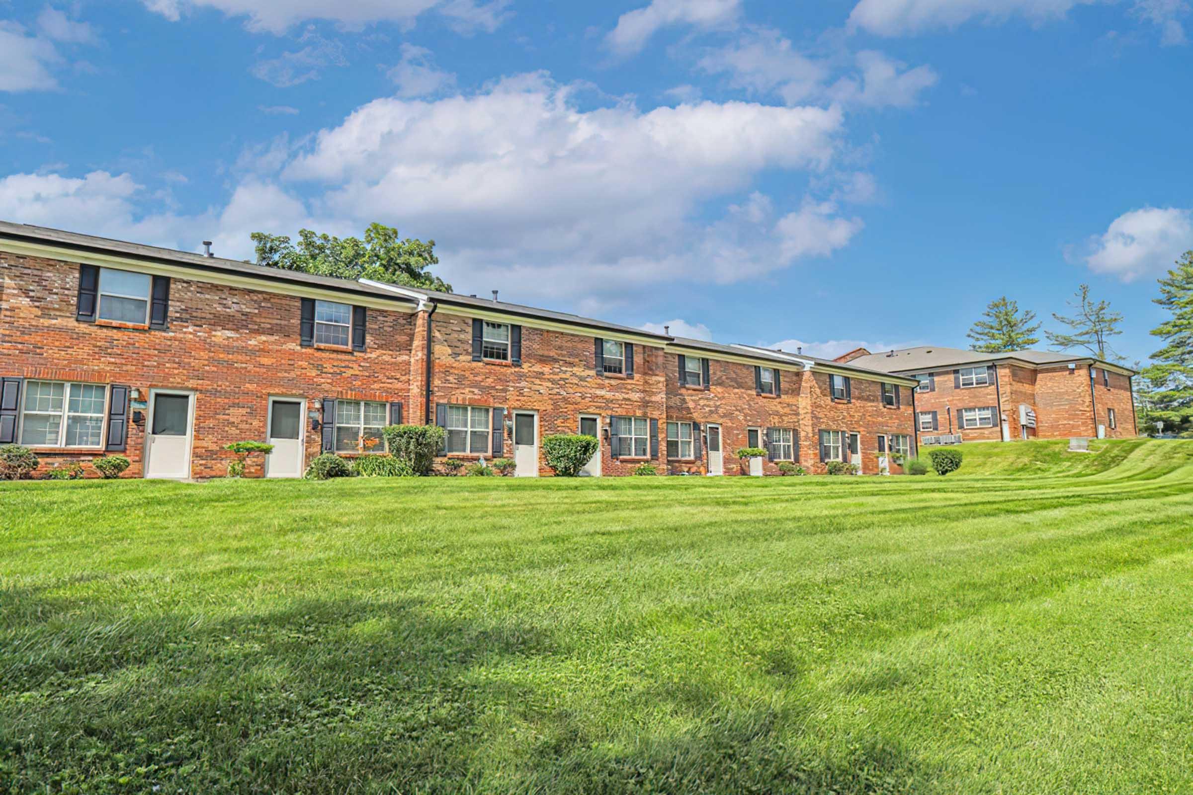 A row of brick apartment buildings set against a clear blue sky. Green grass and landscaped bushes in the foreground create a pleasant outdoor space. The buildings feature multiple windows and entrance doors, demonstrating a well-maintained residential area.