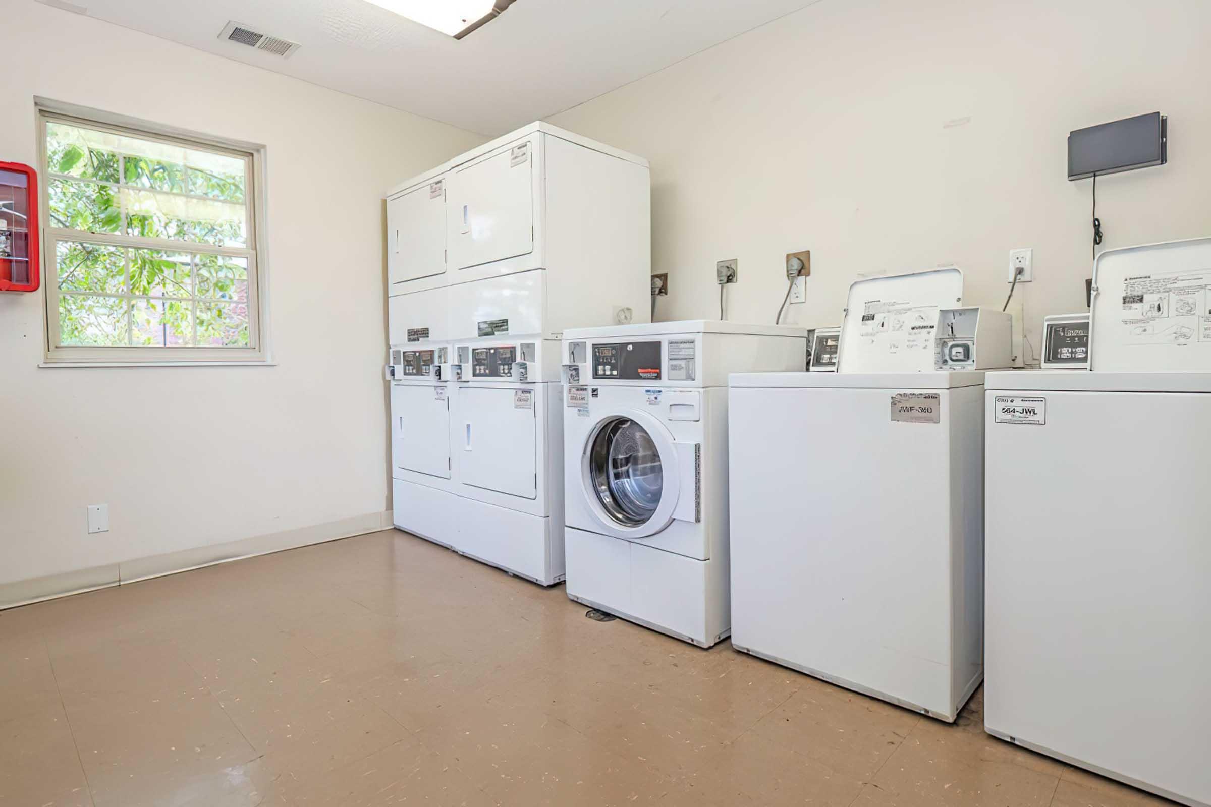 A laundry room featuring multiple coin-operated washing machines and dryers, with a window allowing natural light. The machines are white, arranged neatly against the wall, and the floor is a light brown. A small wall-mounted monitor is visible, along with a fire extinguisher on the wall.