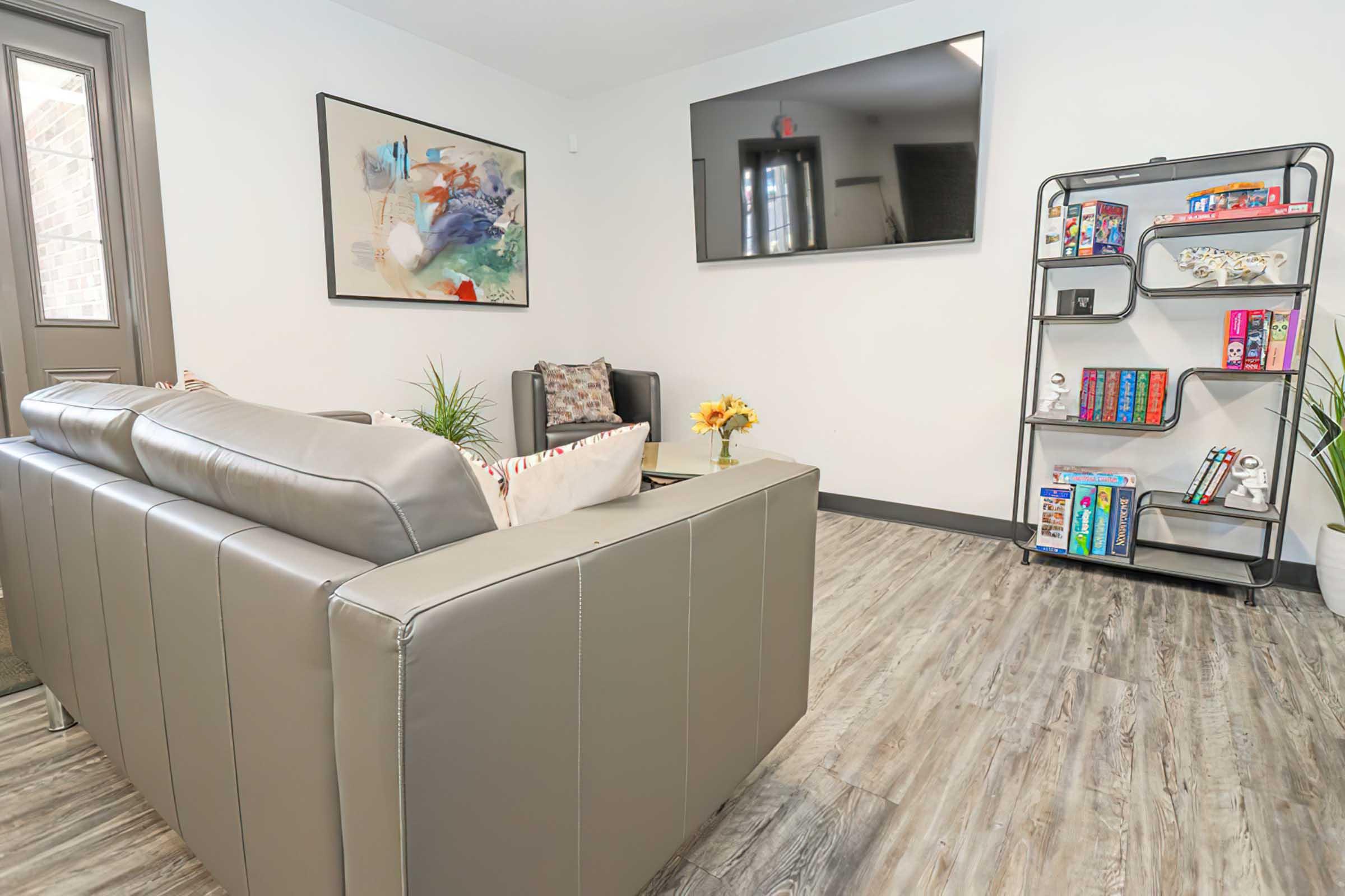 A modern living space featuring a gray sofa with decorative pillows, a colorful abstract painting on the wall, and a flat-screen TV. There's a cozy reading nook with plants and a small bookshelf filled with books and decorative items. The wood-style flooring adds warmth to the room.