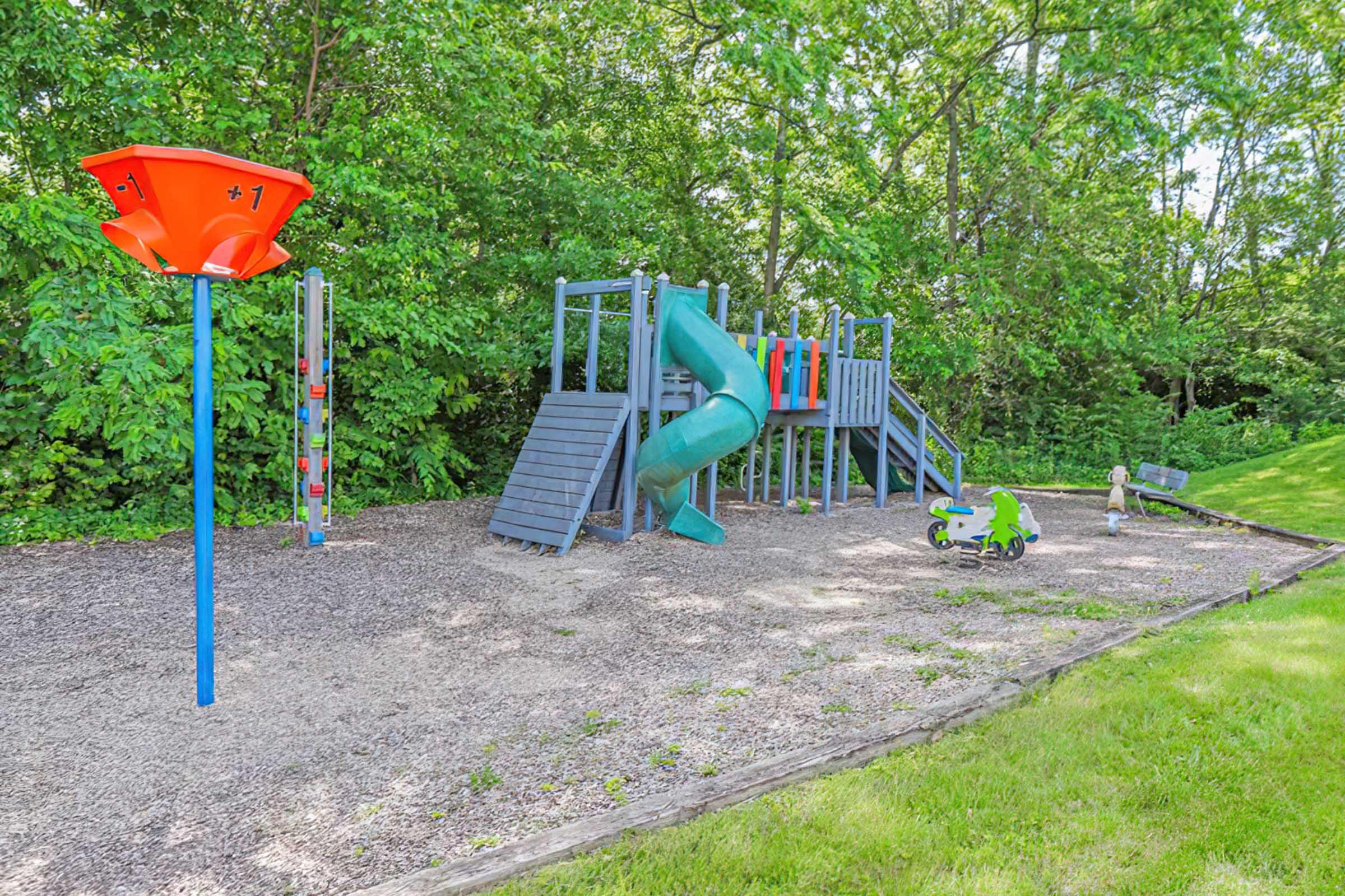 A playground featuring a colorful slide, climbing structure, and a water play feature. There is a small green tricycle on the ground and the surrounding area is covered with mulch and grass, with trees providing a lush green backdrop. The scene is bright and inviting, ideal for children to play.