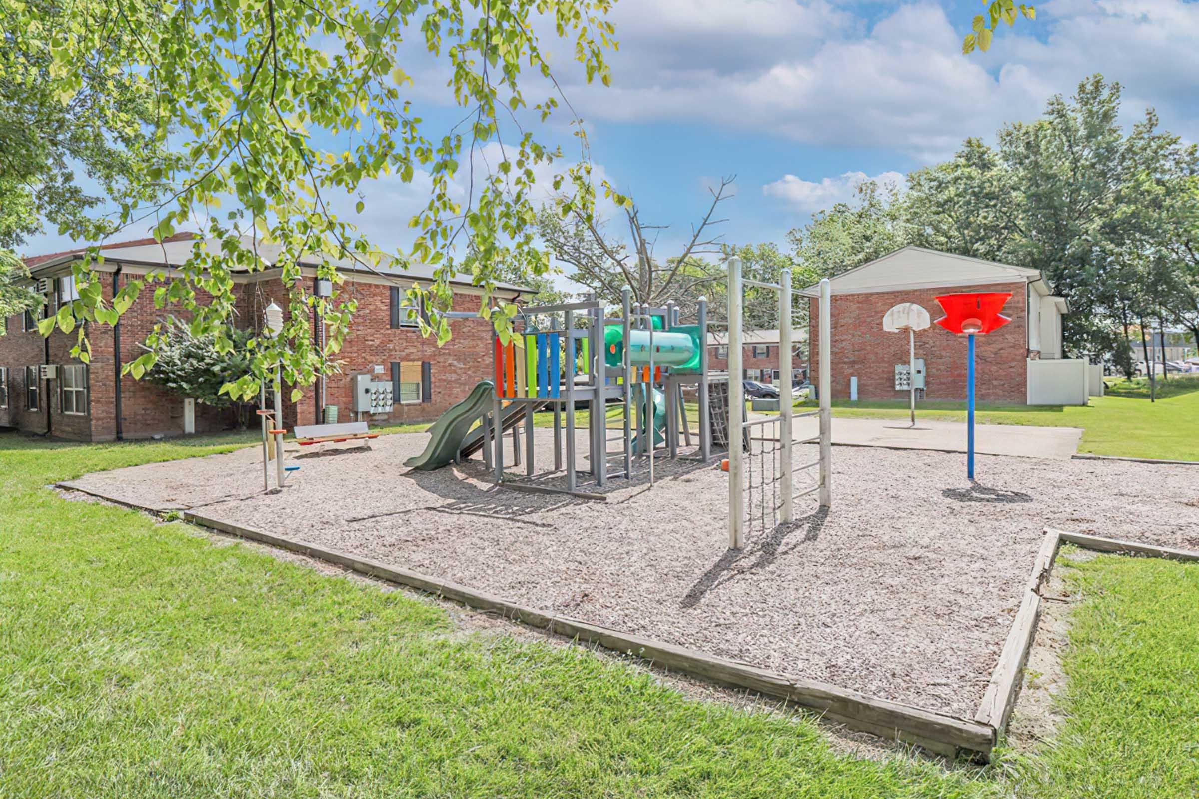 A playground area featuring a slide, climbing structure, and basketball hoop, surrounded by grass and trees. In the background, there are brick apartment buildings, and the sky is clear with a few clouds. The playground surface consists of wood chips for safety.