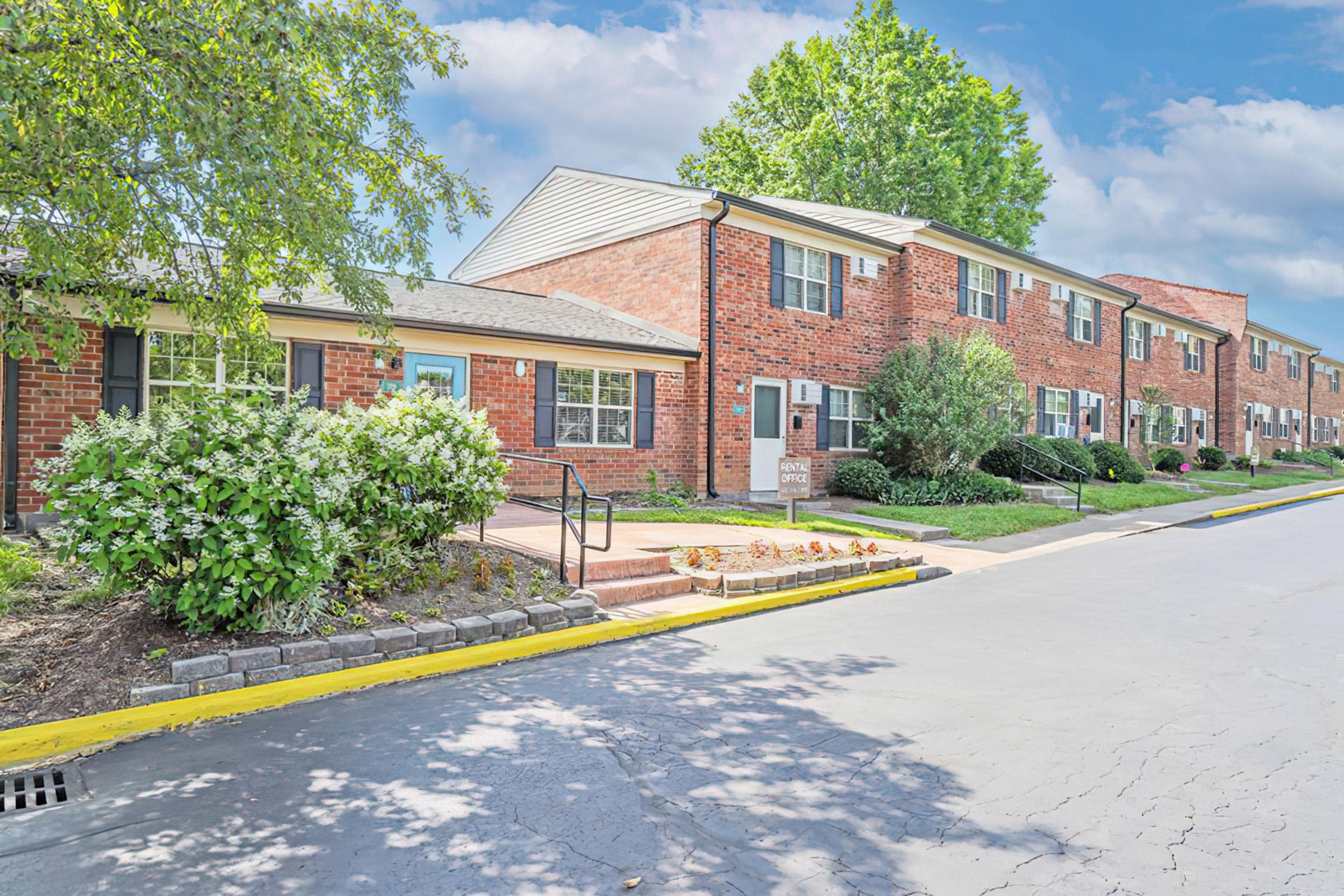 A well-maintained brick apartment complex with landscaped gardens. The entrance features a pathway lined with bushes and small trees, and there are several units visible, each with windows and outdoor space. A clear blue sky is in the background, creating a bright and inviting atmosphere.