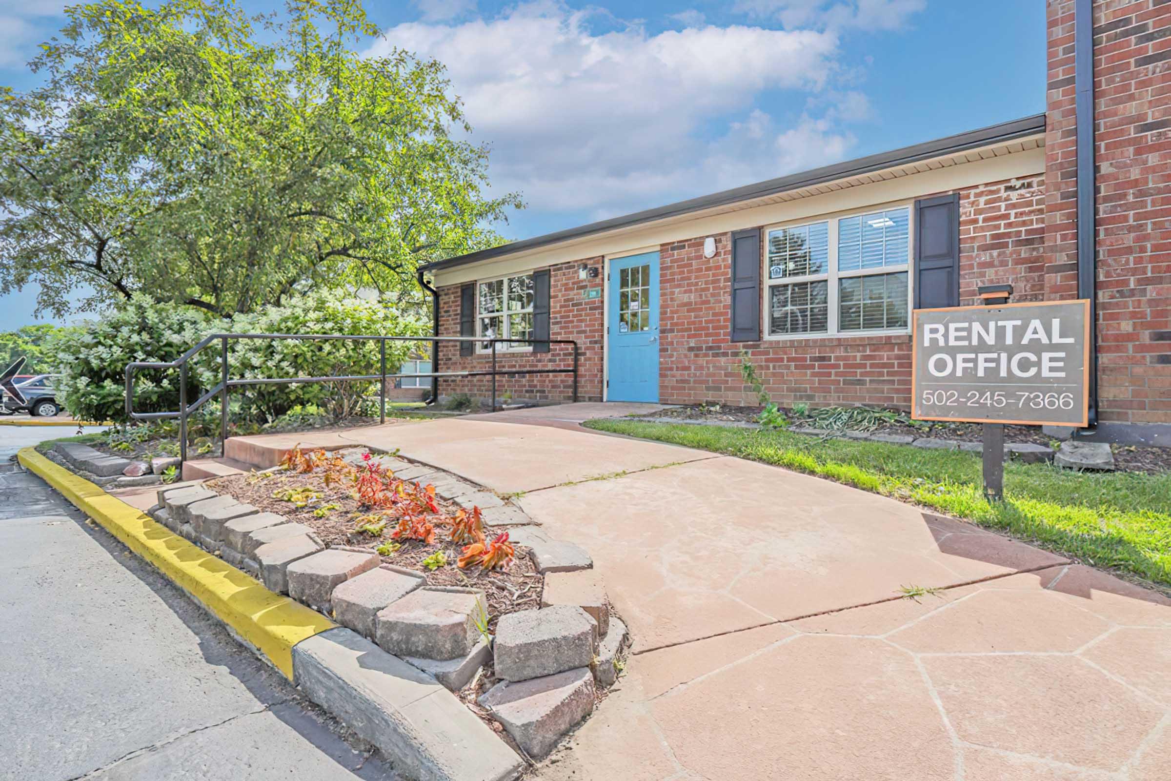 A view of a rental office building with a blue door, surrounded by greenery. A sign in front reads "RENTAL OFFICE" along with a phone number. The entrance features a paved walkway with flowerbeds on the side, providing a welcoming atmosphere. Bright blue sky and fluffy clouds are visible in the background.