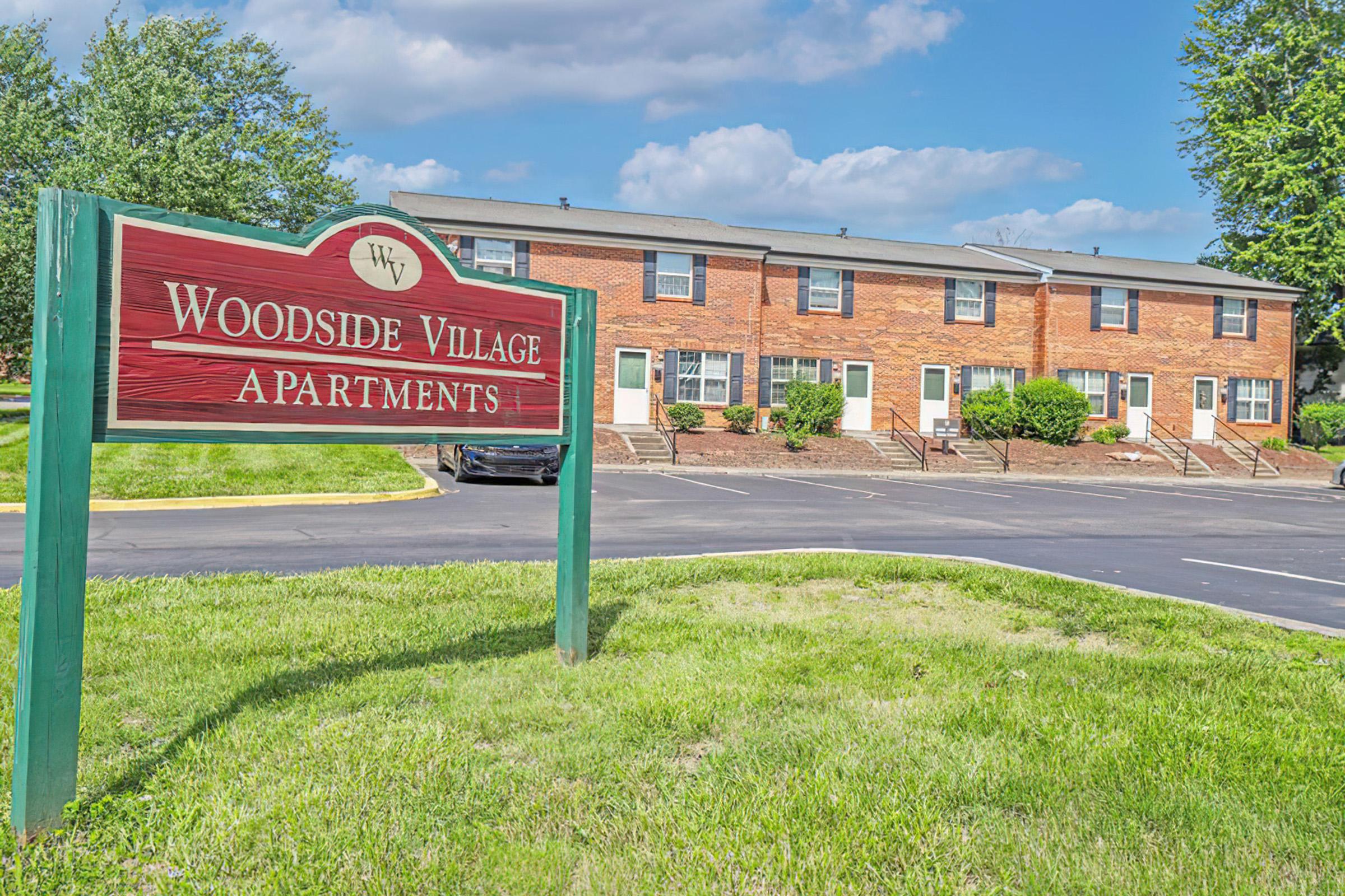 Sign for Woodside Village Apartments in front of a two-story brick building complex. The area is landscaped with green grass and bushes under a clear blue sky with a few clouds. There is a parking area visible in front of the apartments.