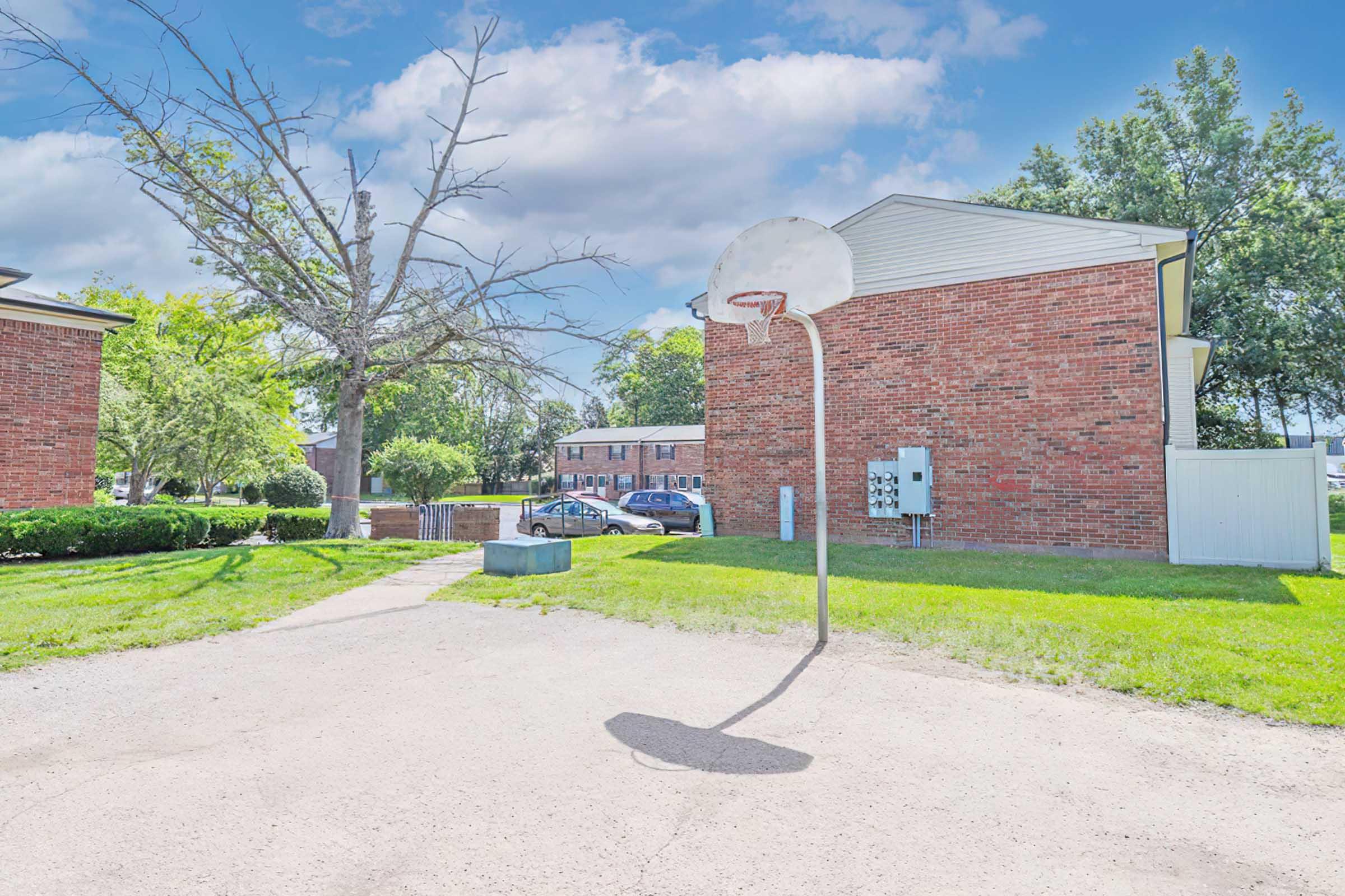 A sunny outdoor scene featuring a basketball hoop with a shadow on a gravel surface. In the background, there are two brick buildings and green grass, along with a few trees and parked cars. The setting appears to be a residential area.