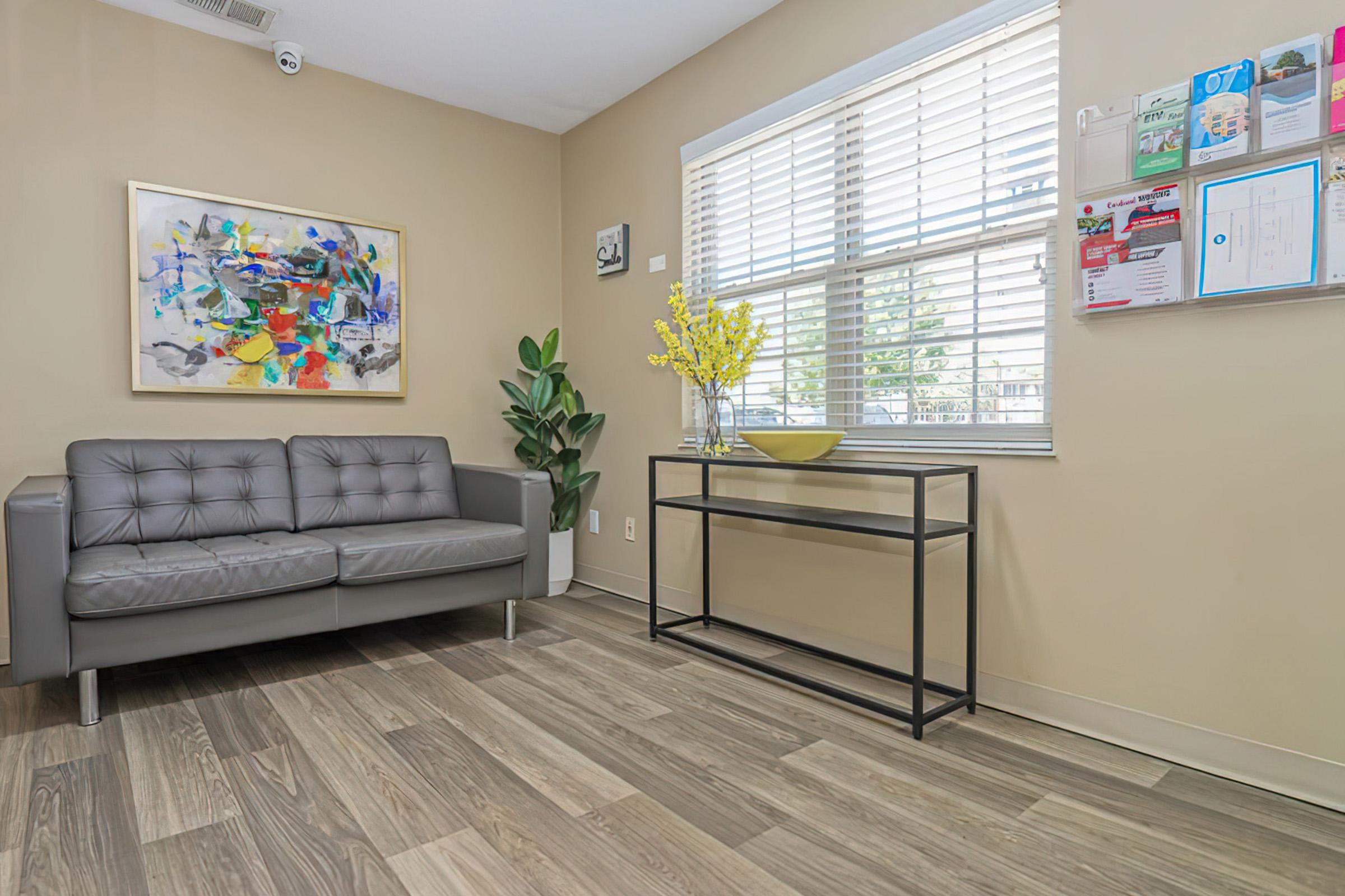 A modern waiting room featuring a gray leather couch, a minimalist black metal table with a yellow vase, and bright yellow flowers. A wall-mounted display holds various brochures and notifications. Large windows provide natural light, and the walls are painted a warm beige color, creating a welcoming atmosphere.
