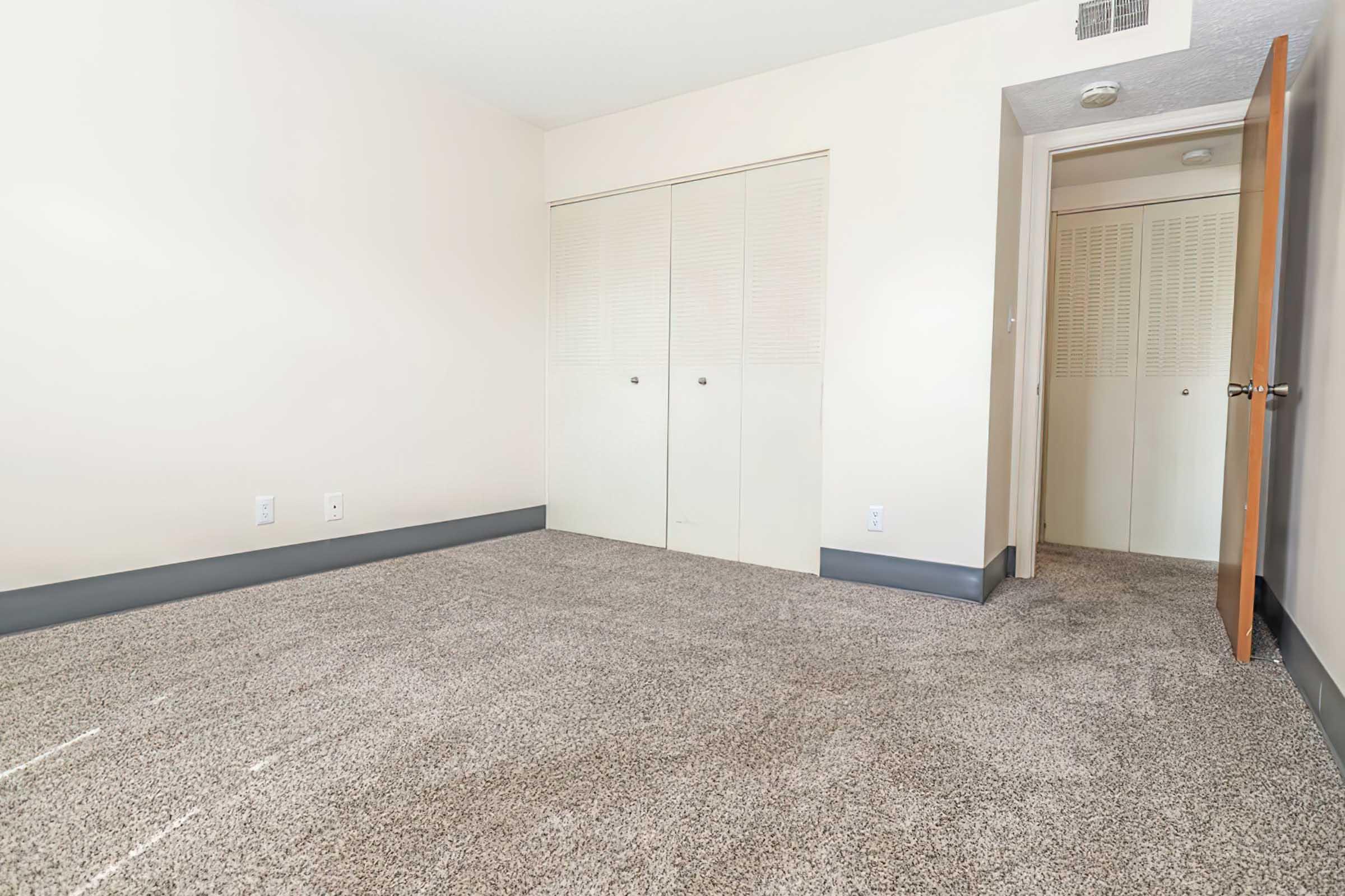Empty room with beige walls and light-colored carpet. There are two closet doors on one wall and an open doorway leading to another space. The room is well-lit with natural light and has minimal furnishings.