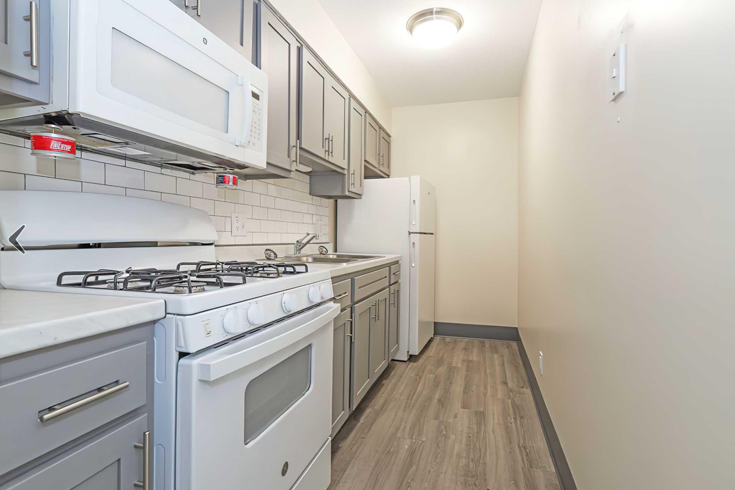 A modern kitchen featuring gray cabinets, a white gas stove, a microwave above, and a refrigerator. The countertops are light-colored, and the floor is a wood-like laminate. The walls are painted a soft beige, creating a warm, inviting atmosphere.
