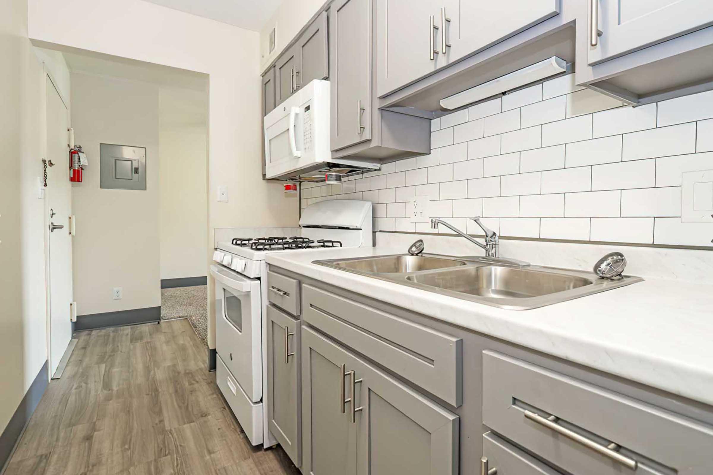 A modern kitchen featuring gray cabinets, a white marble countertop, a stainless steel sink with dual basins, a gas stove, and a microwave. The backsplash consists of white subway tiles. A doorway in the background leads to another room, and there's a small fire safety notice on the wall. The flooring is a wood-like laminate.