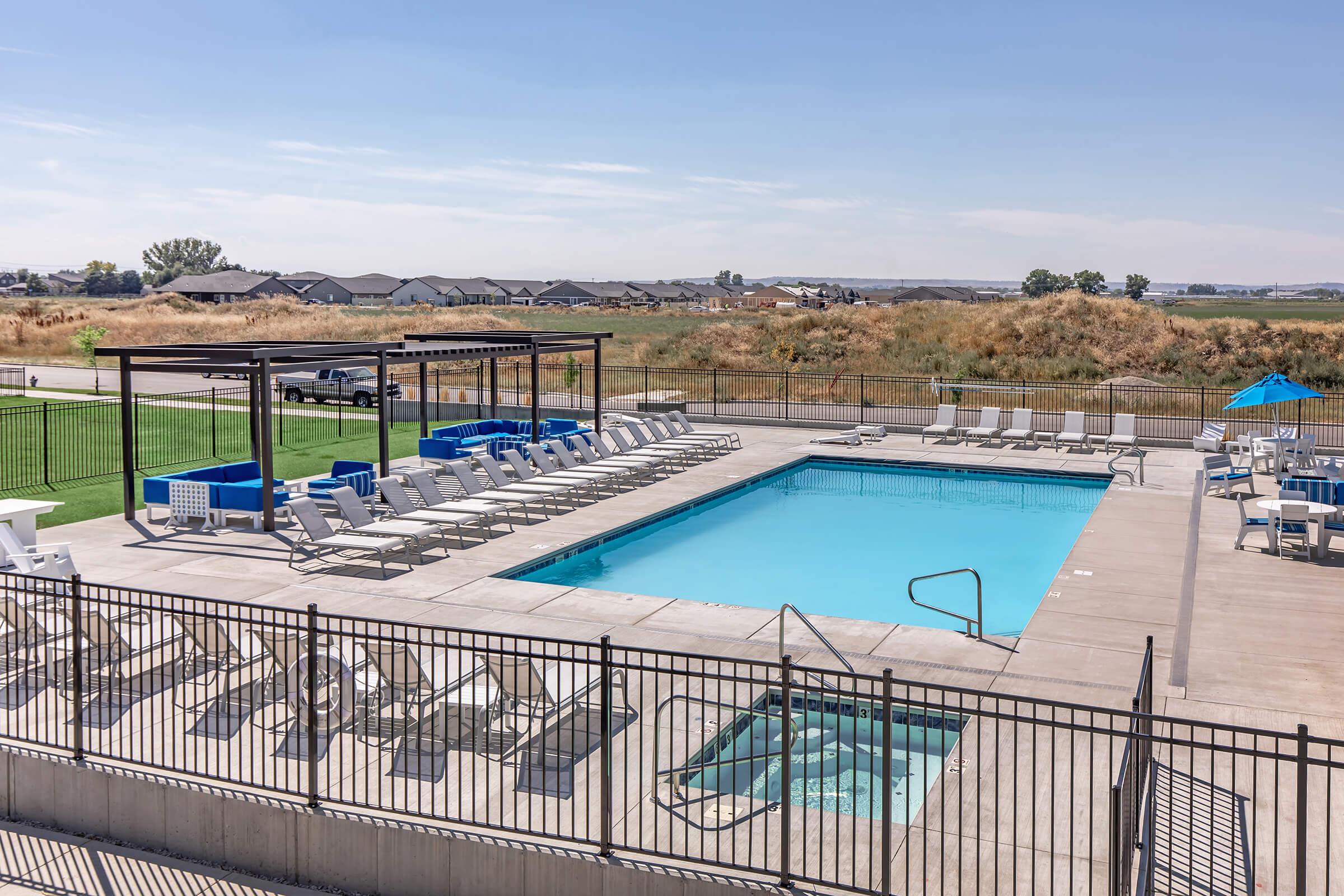 A sunny outdoor pool area featuring a rectangular swimming pool surrounded by lounge chairs. There are shaded seating areas with canopies and a hot tub nearby. Lush grass and a view of distant homes are visible in the background under a clear blue sky.