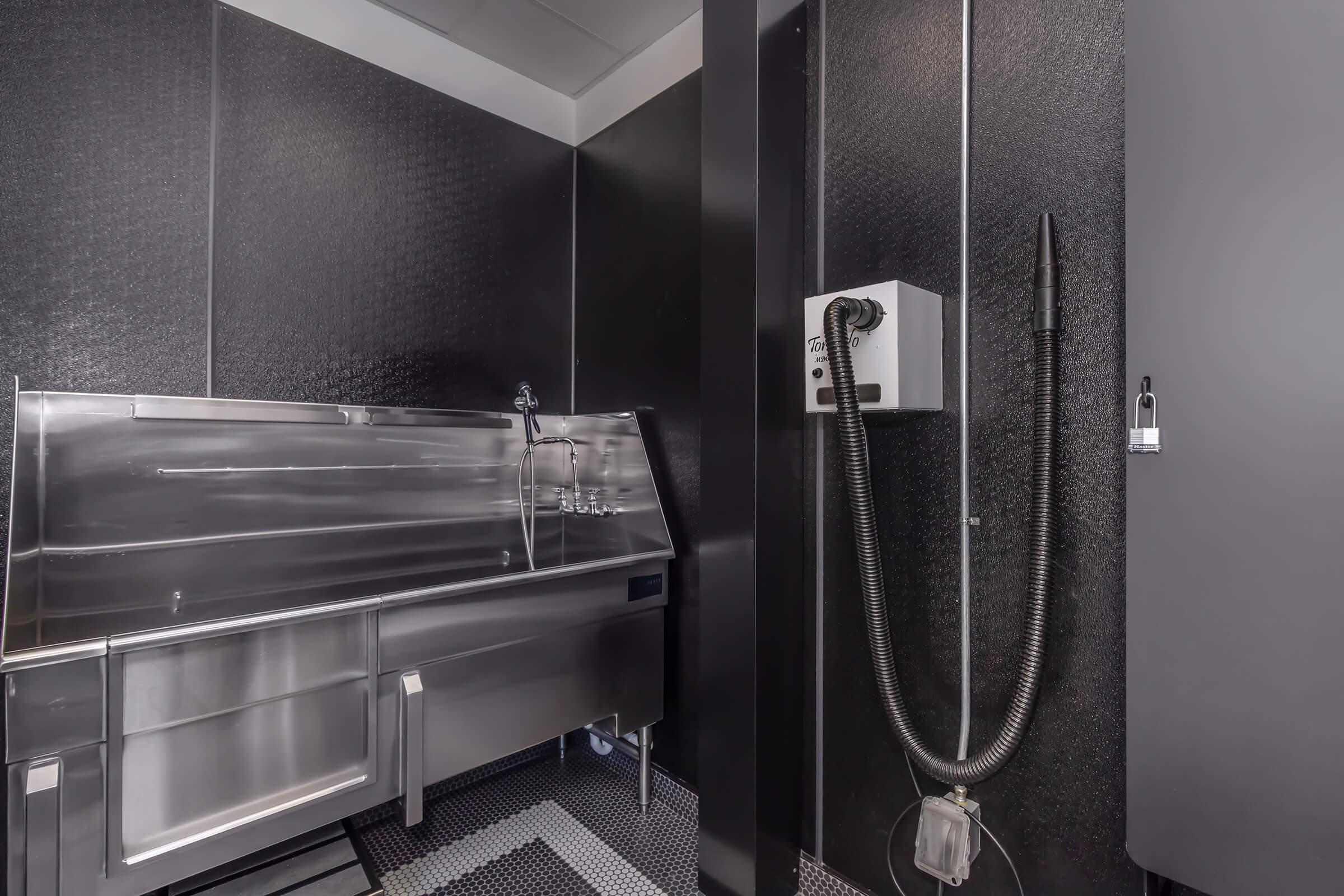A modern stainless steel wash station with a large sink and a handheld sprayer, set against a sleek black textured wall. The area is well-lit and includes a mounted hand dryer. The floor features a checkered pattern, contributing to a clean, utilitarian design suitable for a grooming or veterinary space.