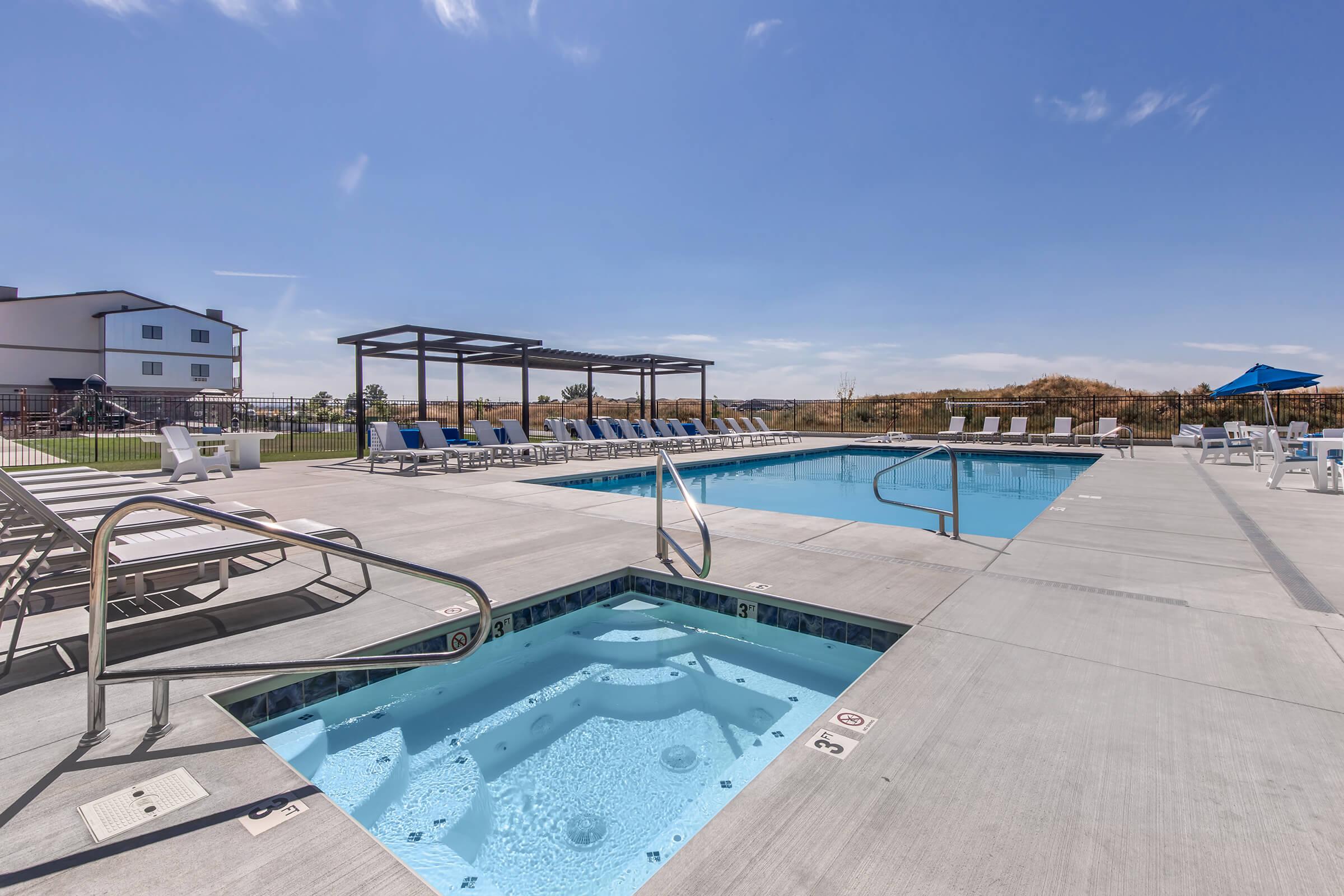 Modern outdoor swimming pool area featuring a clear blue pool, a hot tub, and multiple lounge chairs arranged around the deck. A shaded pergola is visible in the background, along with a spacious lawn and a bright blue sky.