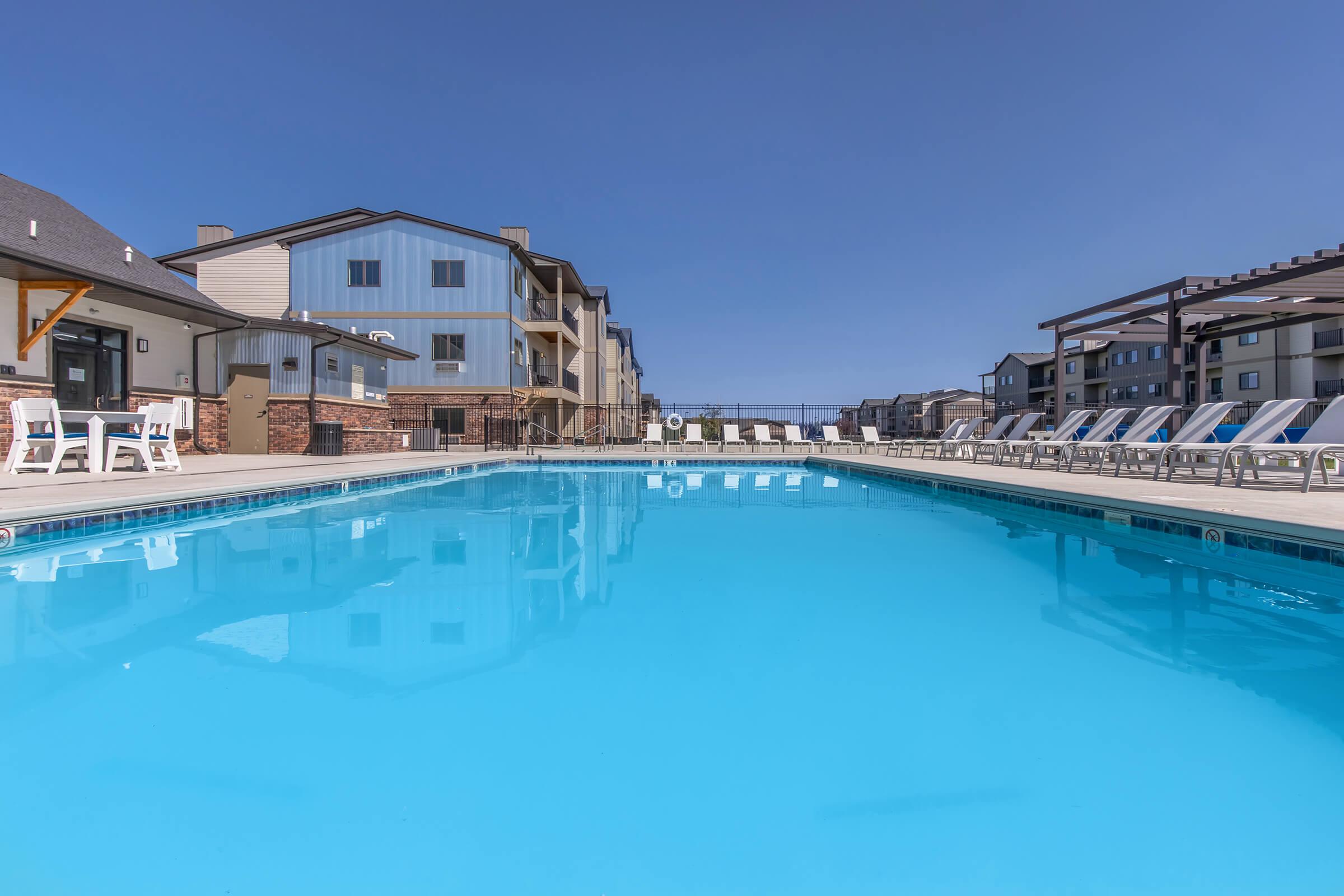 A clear blue swimming pool surrounded by lounge chairs and modern buildings under a bright blue sky. The scene conveys a serene and inviting atmosphere, ideal for relaxation and leisure. The pool reflects the surrounding architecture, adding to the visual appeal of the area.