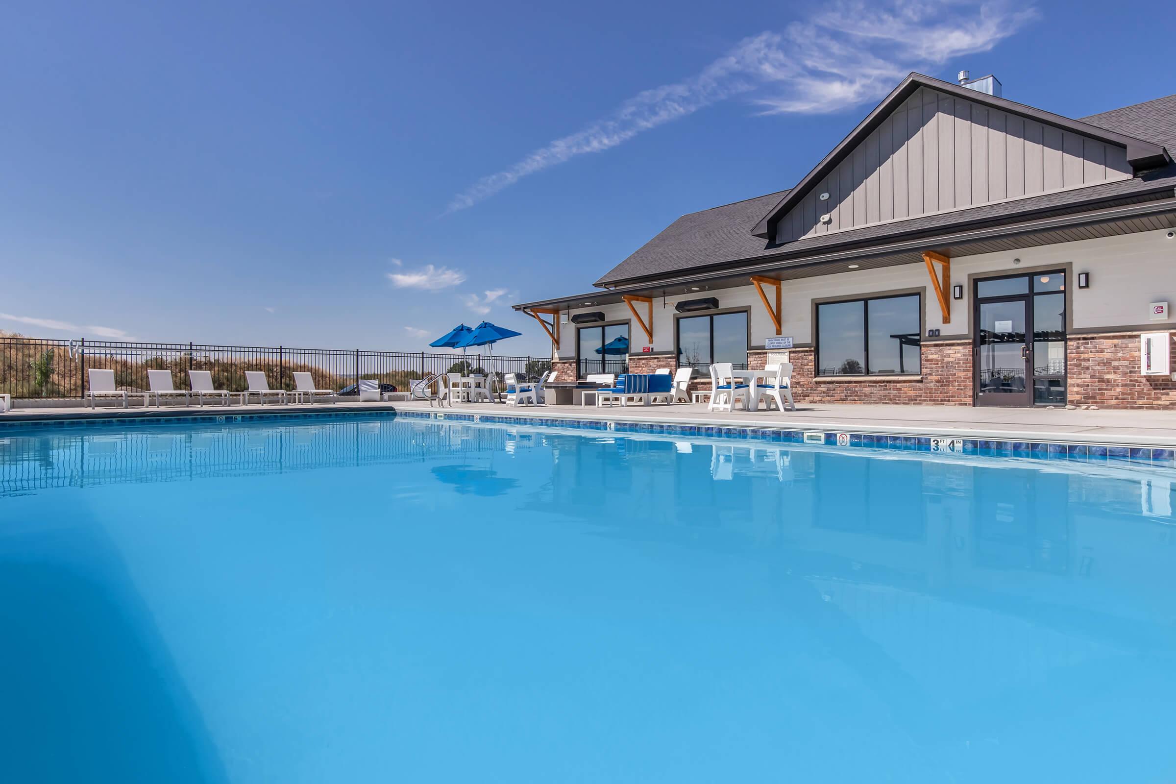 A clear blue swimming pool reflecting the sky, surrounded by a spacious deck with white lounge chairs and an umbrella. In the background, a modern building features large windows and a gray roof, set against a sunny sky with a few clouds. The scene conveys a relaxing outdoor atmosphere.