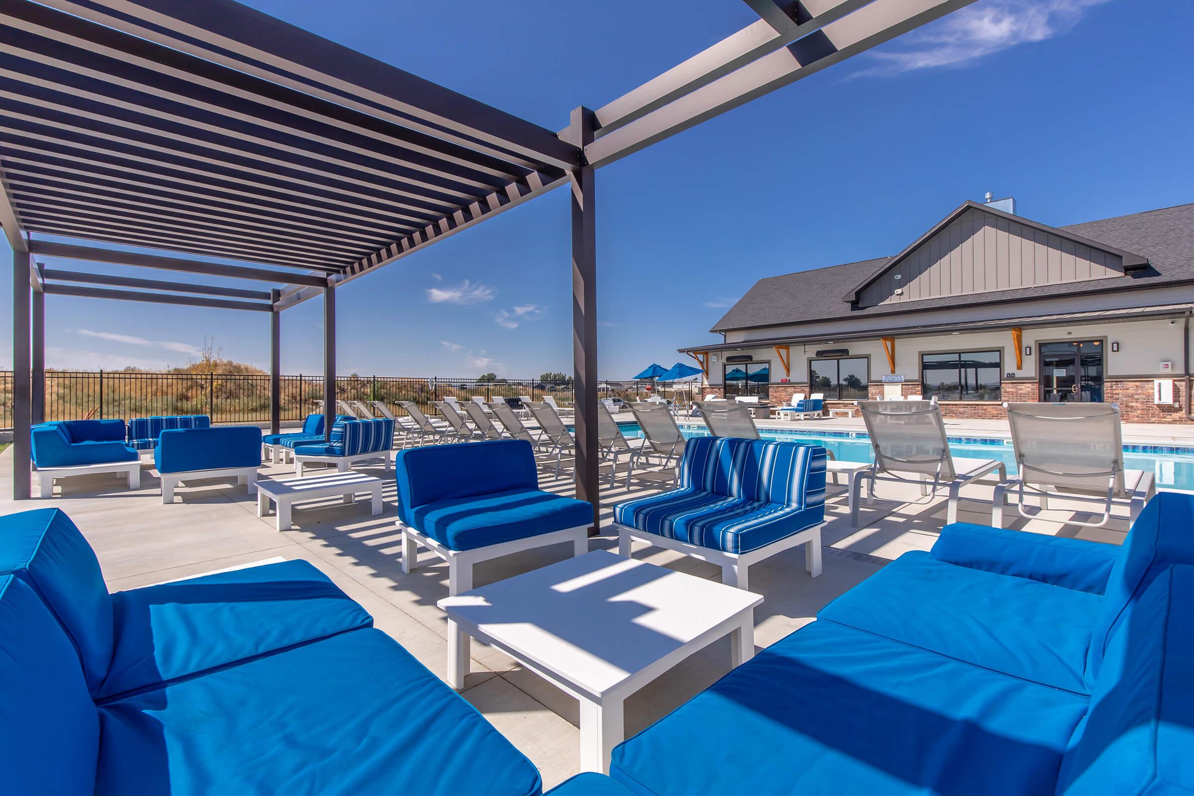 A modern outdoor lounge area featuring bright blue furniture, including comfortable seating and tables. There is a swimming pool in the background, surrounded by sun loungers and a clear blue sky. The design is stylish and inviting, perfect for relaxation and leisure.