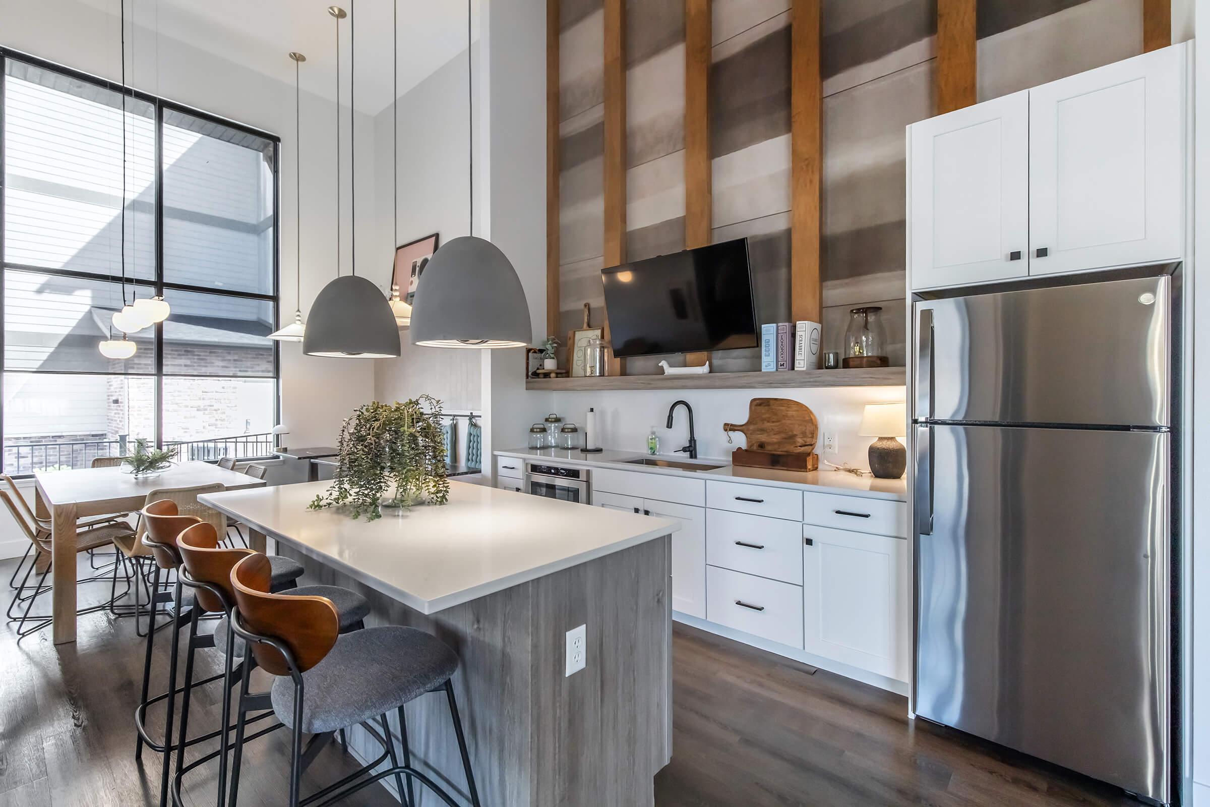Modern kitchen featuring a large island with bar stools, sleek cabinetry, and stainless steel appliances. Large windows provide natural light, and decorative elements include pendant lights and a wall with wooden accents. Dining area visible in the background with a stylish table and chairs.