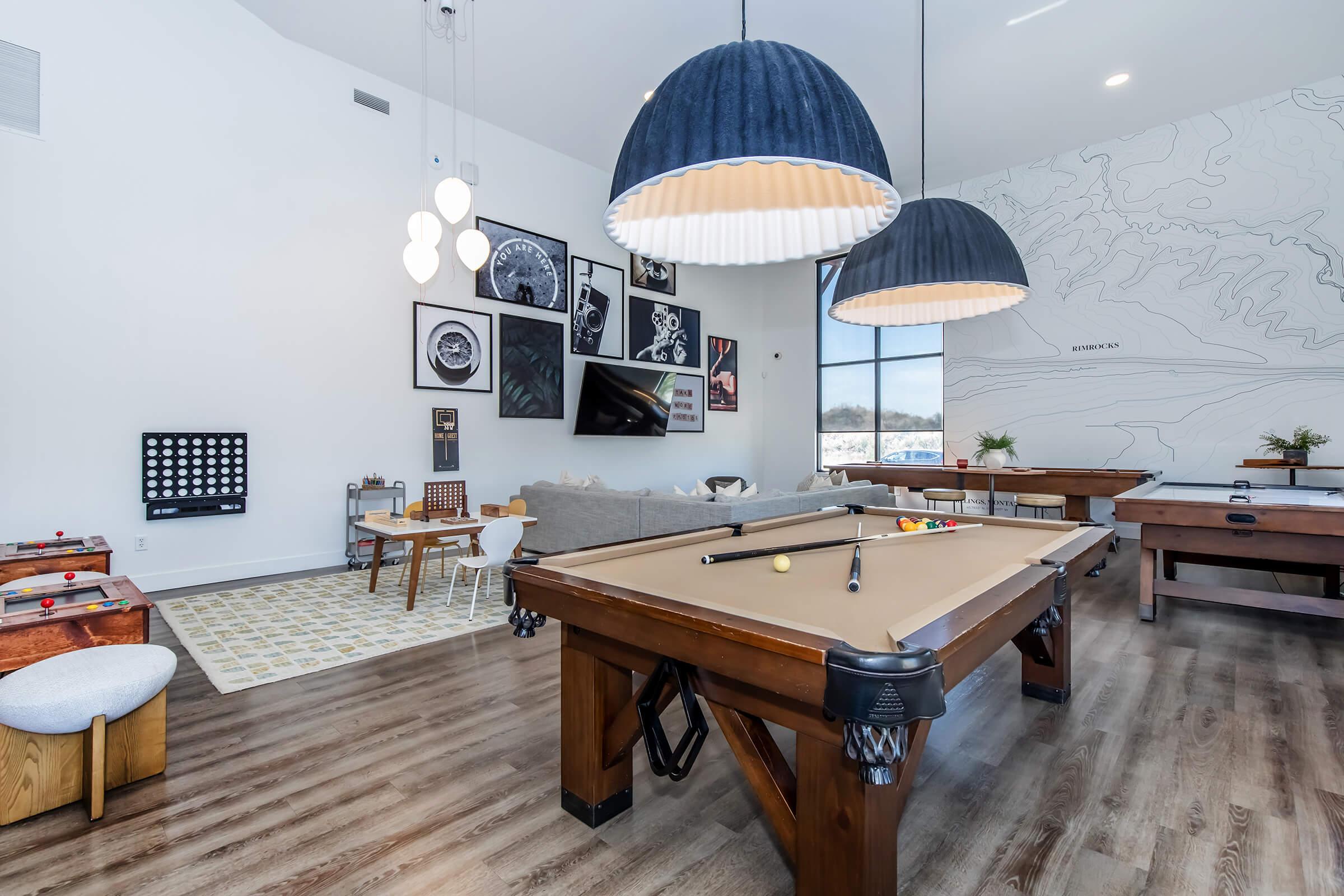 A modern recreational room featuring a pool table, large pendant lights, and a cozy seating area with a sofa and dining table. The walls are adorned with framed artwork, and a large window provides natural light, enhancing the inviting atmosphere.