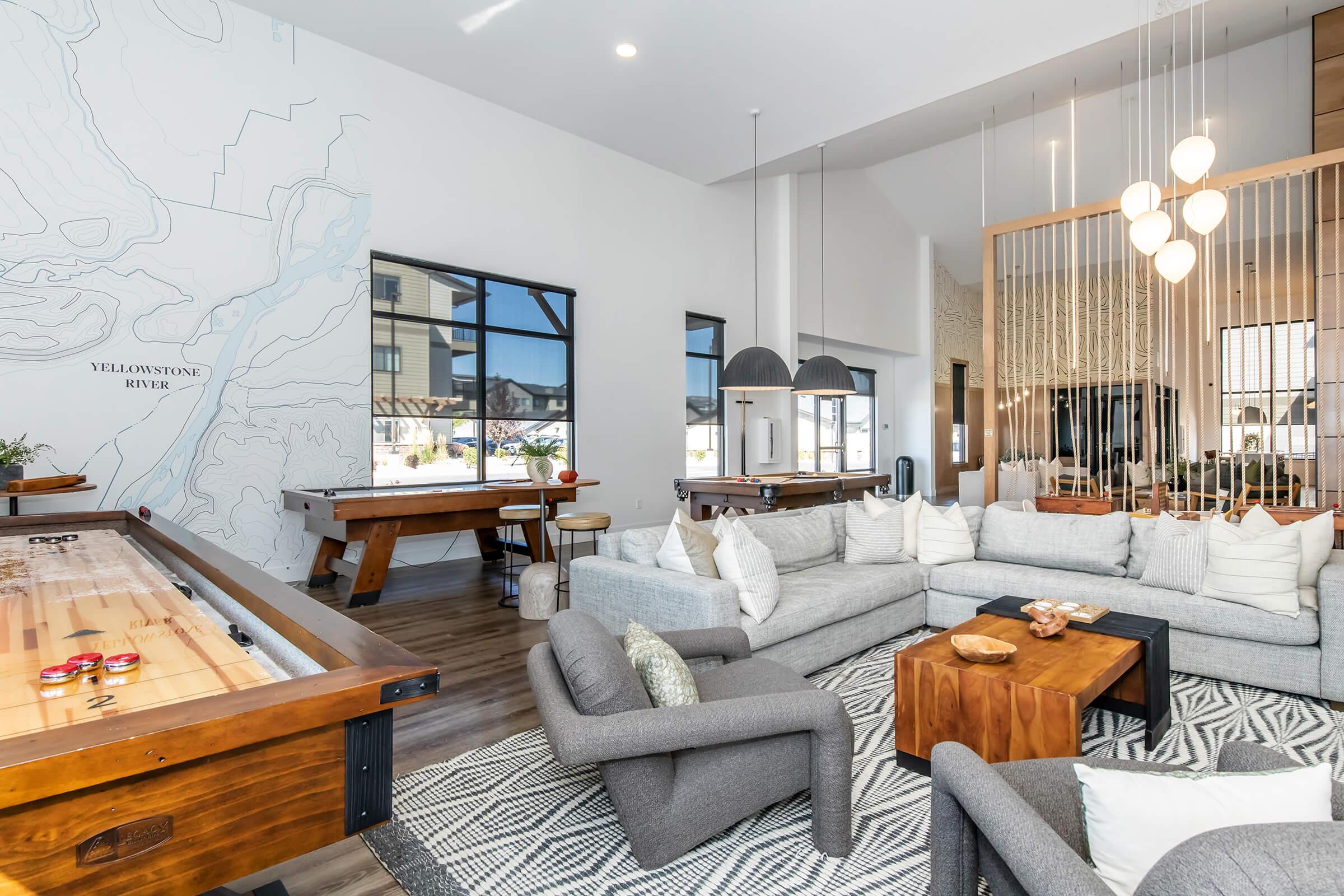 A modern, stylish living area featuring a large grey sectional sofa, a wooden coffee table, and elegant light fixtures. The space includes a pool table, a map mural on the wall, and large windows letting in natural light, creating an inviting atmosphere.