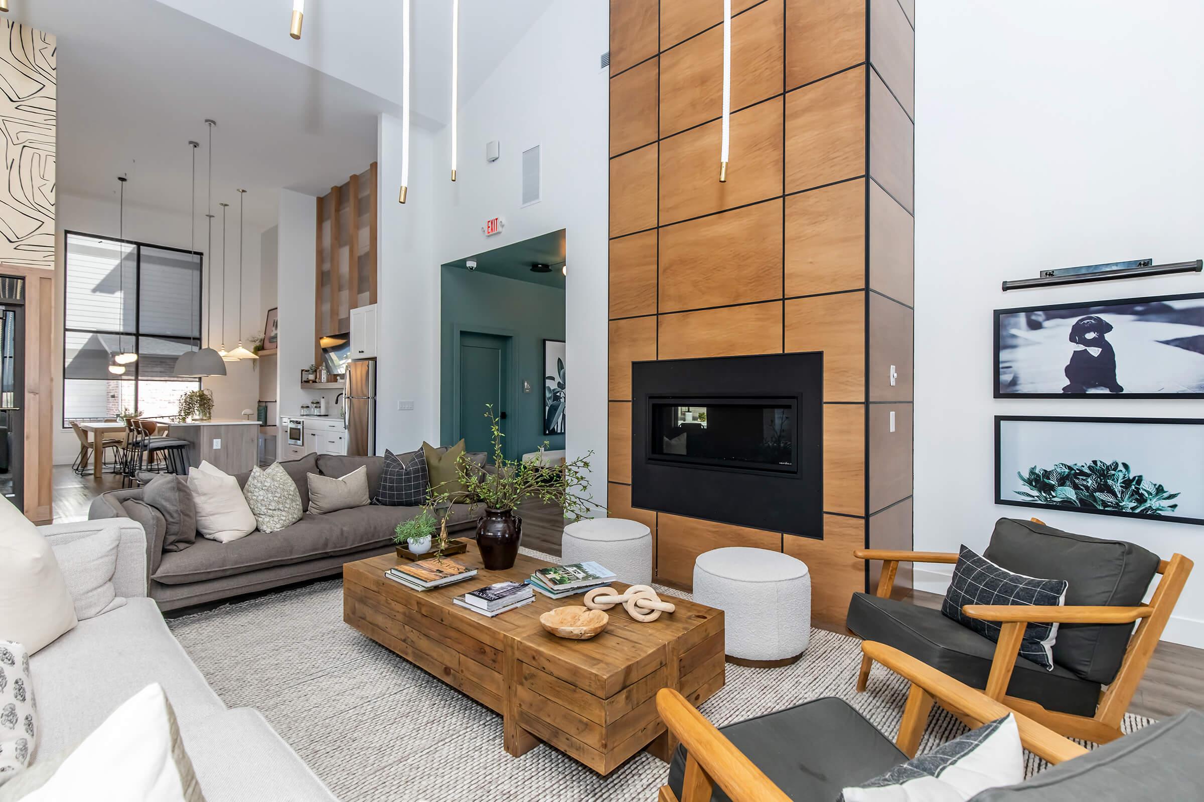 A modern living room featuring a tall wooden accent wall with a fireplace, a large sectional sofa, a wooden coffee table with decorative items, and stylish armchairs. The space is bright and airy, with large windows and contemporary decor, creating a cozy yet elegant atmosphere.