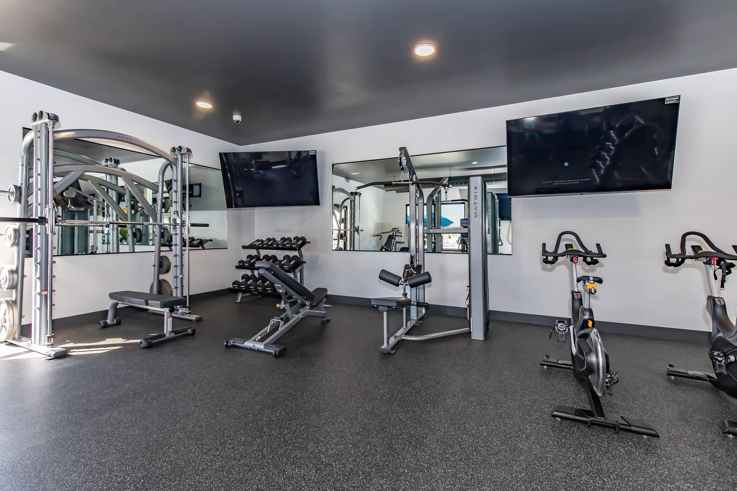 A modern gym interior featuring workout equipment including weight benches, dumbbells, and stationary bikes. There are large wall mirrors and two flat-screen TVs mounted on the walls, all set in a bright, spacious environment with dark flooring.