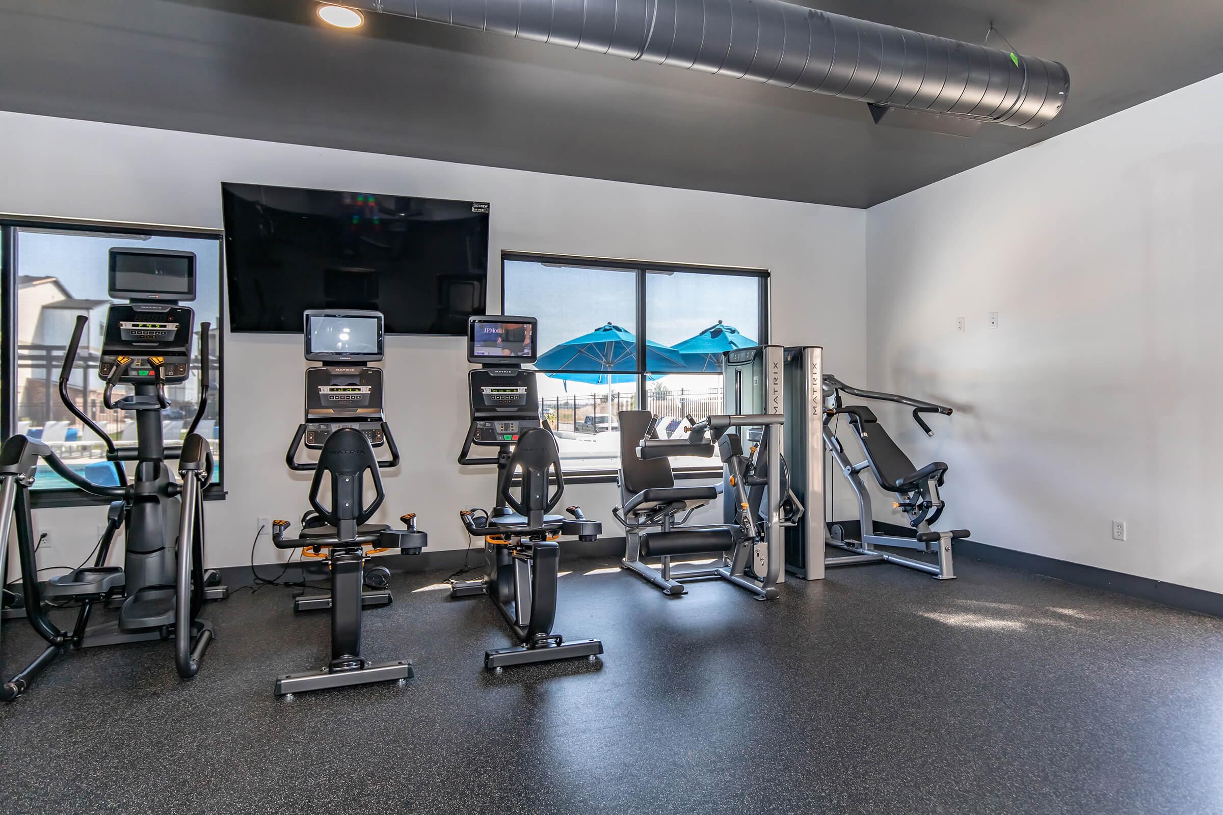 A modern gym interior featuring various exercise equipment, including treadmills, stationary bikes, and weight machines. Large windows let in natural light, showcasing blue umbrellas outside. The space has a sleek, clean design with dark flooring and a minimalistic aesthetic.