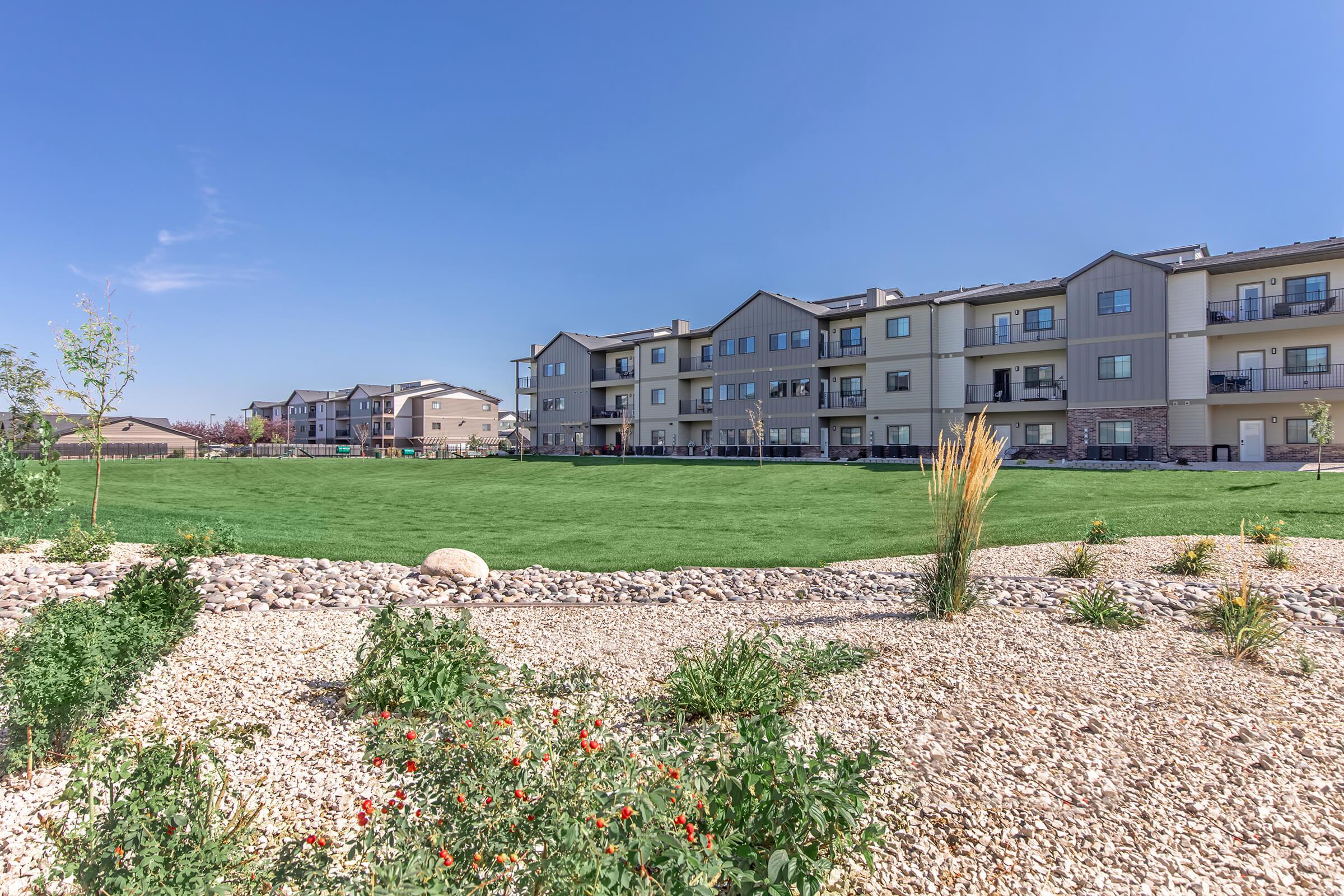 A view of a residential apartment complex set against a clear blue sky. The foreground features landscaped areas with greenery and decorative stones. Multiple buildings are shown in the background, each with balconies, surrounded by a large, well-maintained lawn.