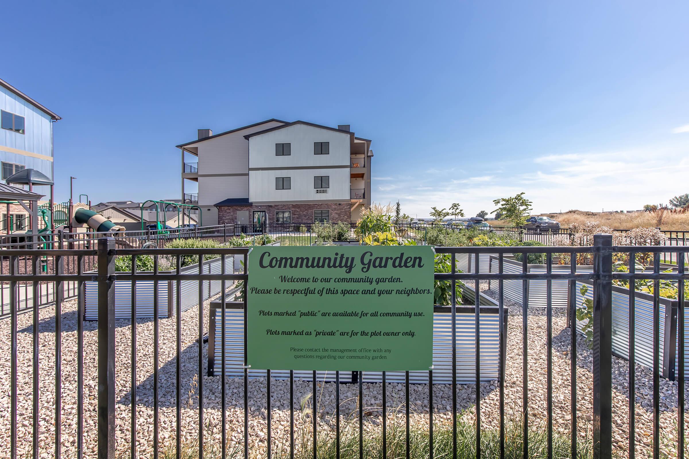 A gated community garden with a sign that reads "Community Garden. Welcome to our community garden. Please be respectful of this space and your neighbors." In the background, there are residential buildings and a playground area. The garden features raised beds and gravel pathways.