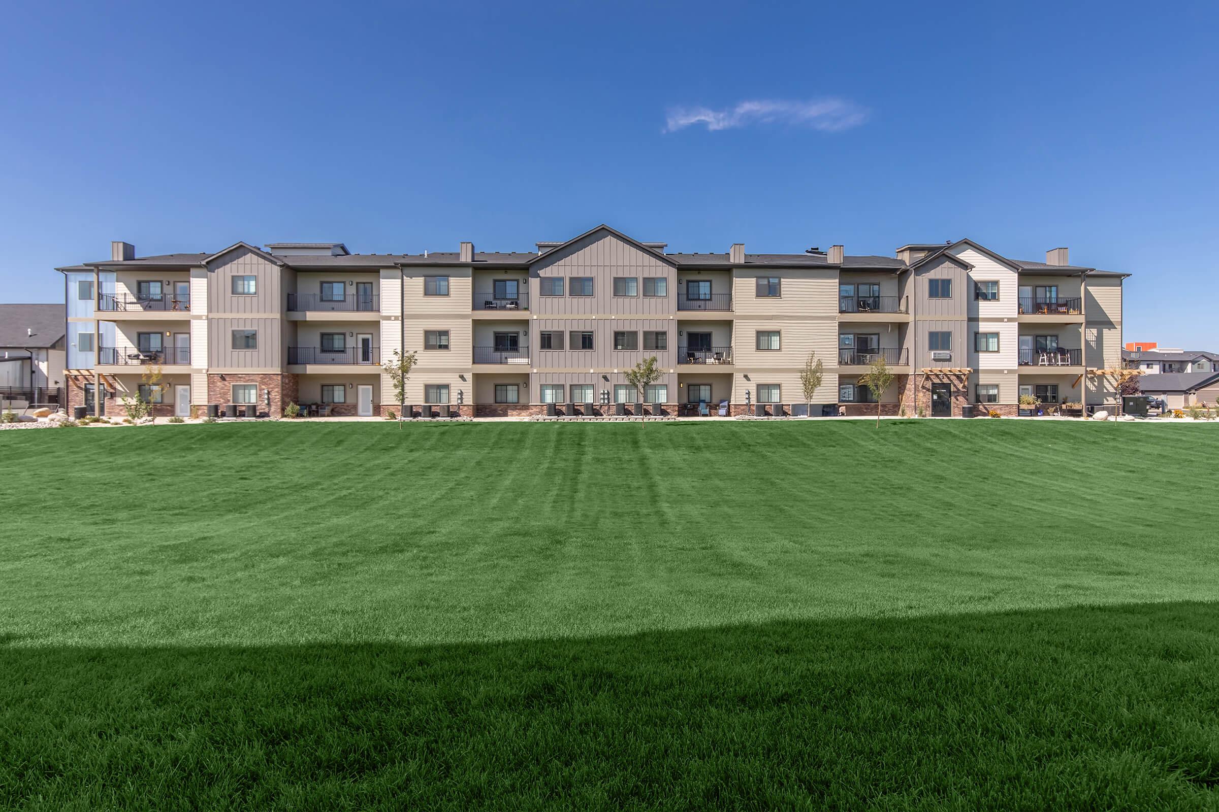 A modern apartment building with multiple units, featuring a mix of light and dark siding. The structure has balconies and a well-maintained lawn in the foreground, under a clear blue sky. The surrounding area is landscaped, contributing to a welcoming residential environment.