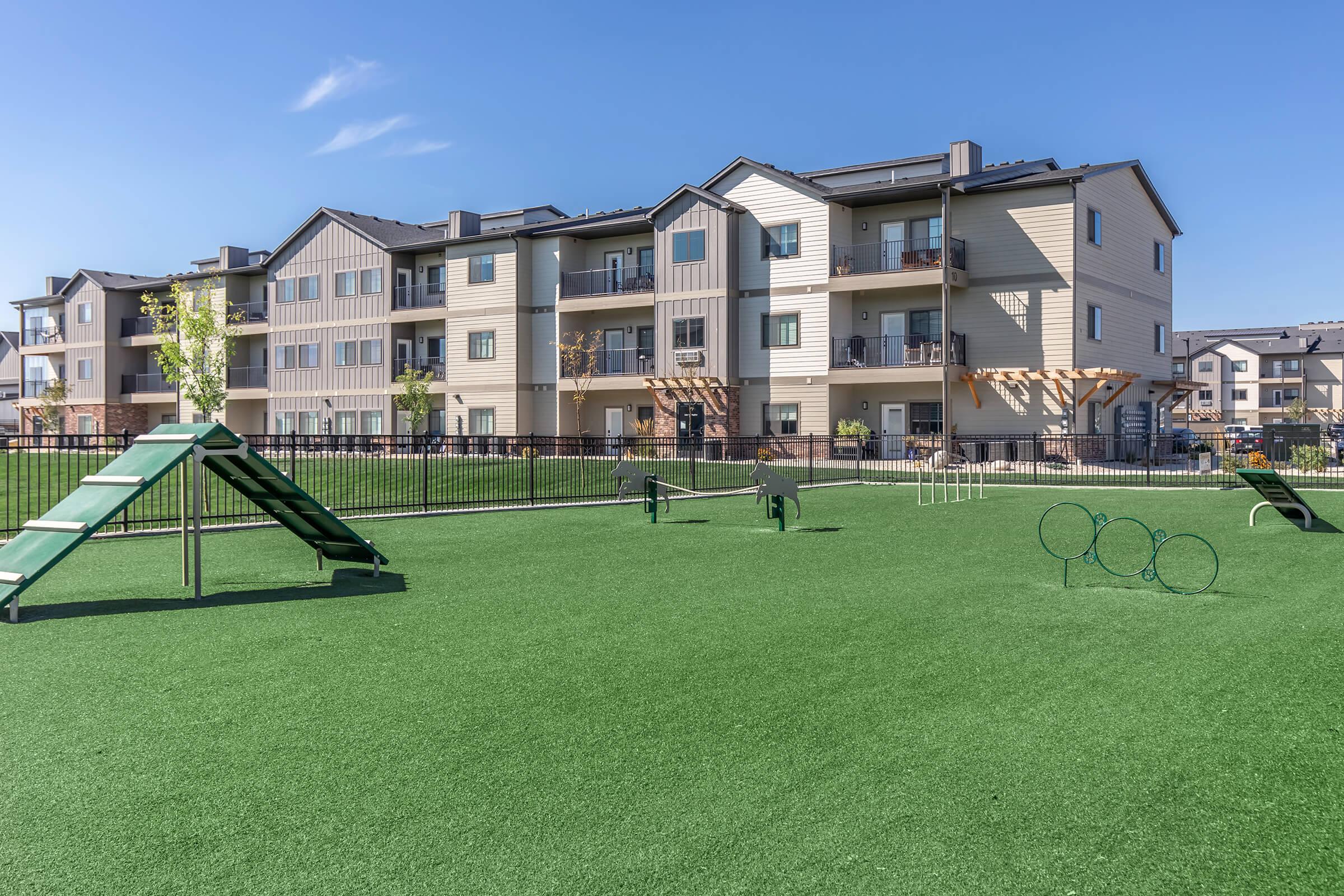 A well-maintained dog park featuring artificial turf, agility equipment like tunnels and hoops, and a fenced area. In the background, there are multi-story residential buildings with balconies under a clear blue sky.