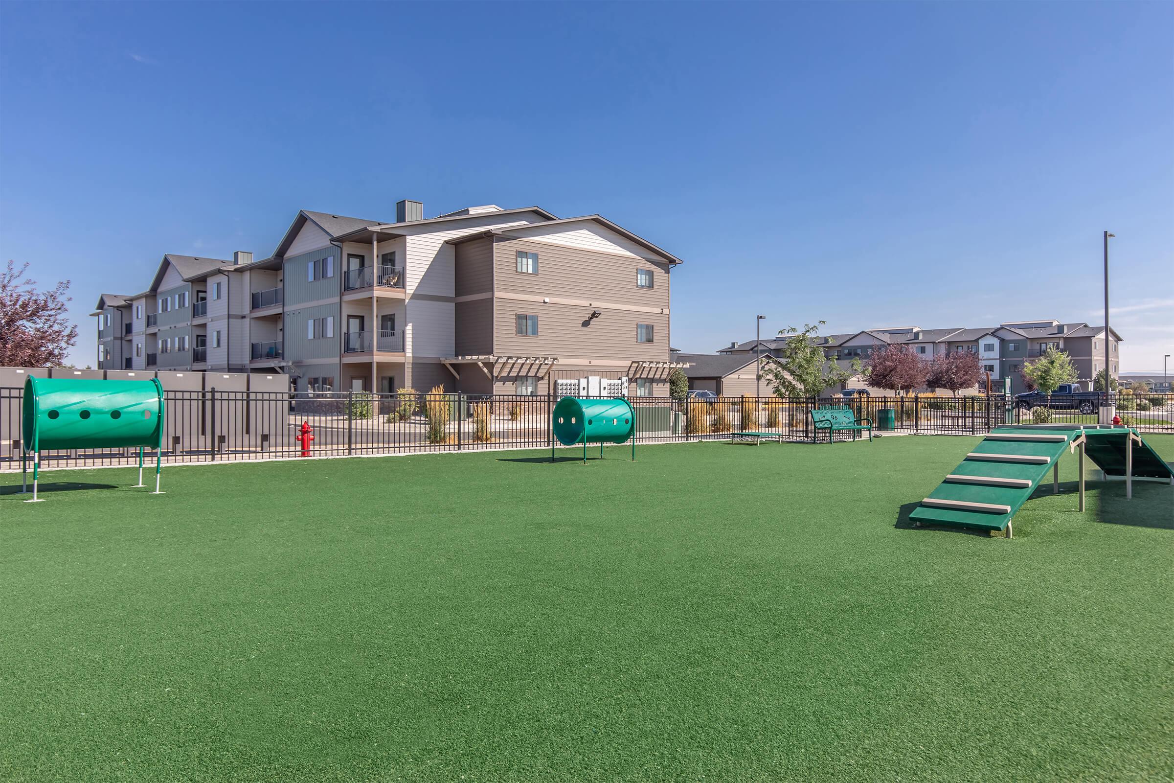 A well-maintained outdoor dog park featuring green artificial turf, agility equipment including tunnels and ramps, and fenced areas. In the background, residential buildings are visible under a clear blue sky, creating a spacious and inviting atmosphere for pets and their owners.