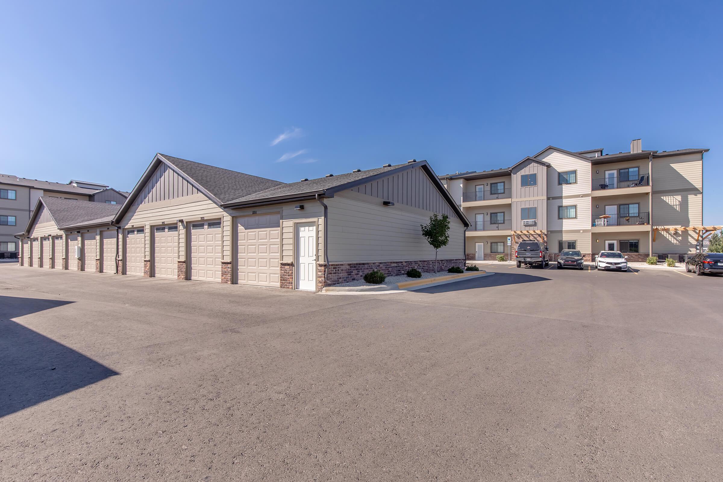A modern apartment complex featuring multiple buildings with a mix of residential units and garage-style storage. The foreground shows paved parking spaces and garages, while the background includes a three-story apartment building with balconies and clear blue skies.