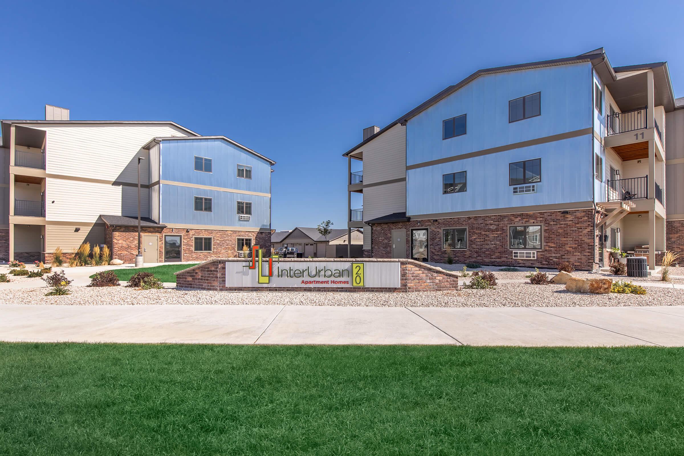 Two modern apartment buildings featuring a mix of blue and beige siding, with a landscaped area and sign for InterUrban Apartments prominently displayed in front. The clear blue sky provides a bright backdrop, and a grassy area lines the pathway leading to the entrance.