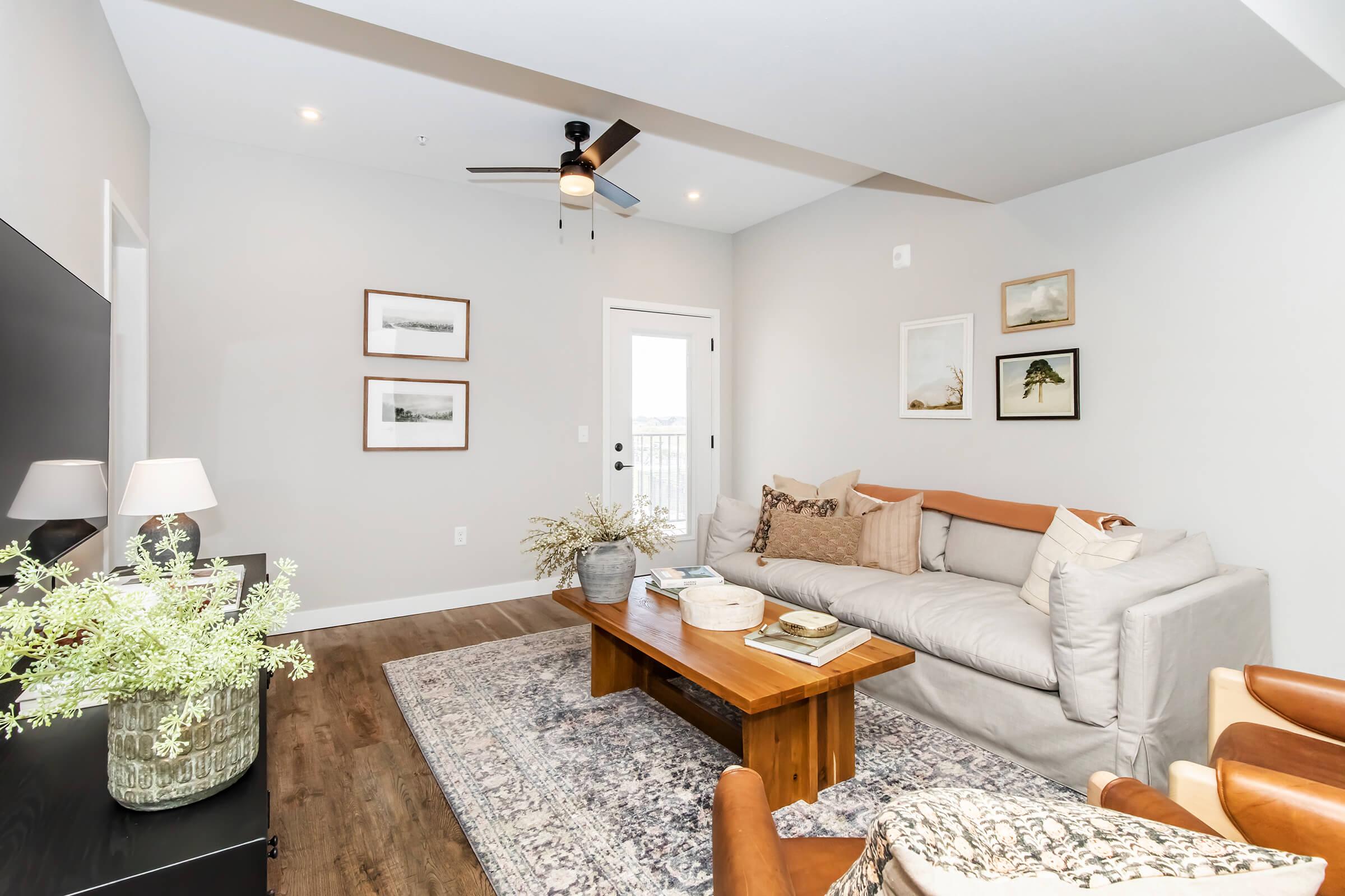 Cozy living room featuring a light gray sectional sofa with decorative pillows, a wooden coffee table, and a patterned area rug. The room has a ceiling fan, light walls, and framed artwork on the walls. A small vase with flowers and a lamp are on the coffee table, enhancing the inviting atmosphere.