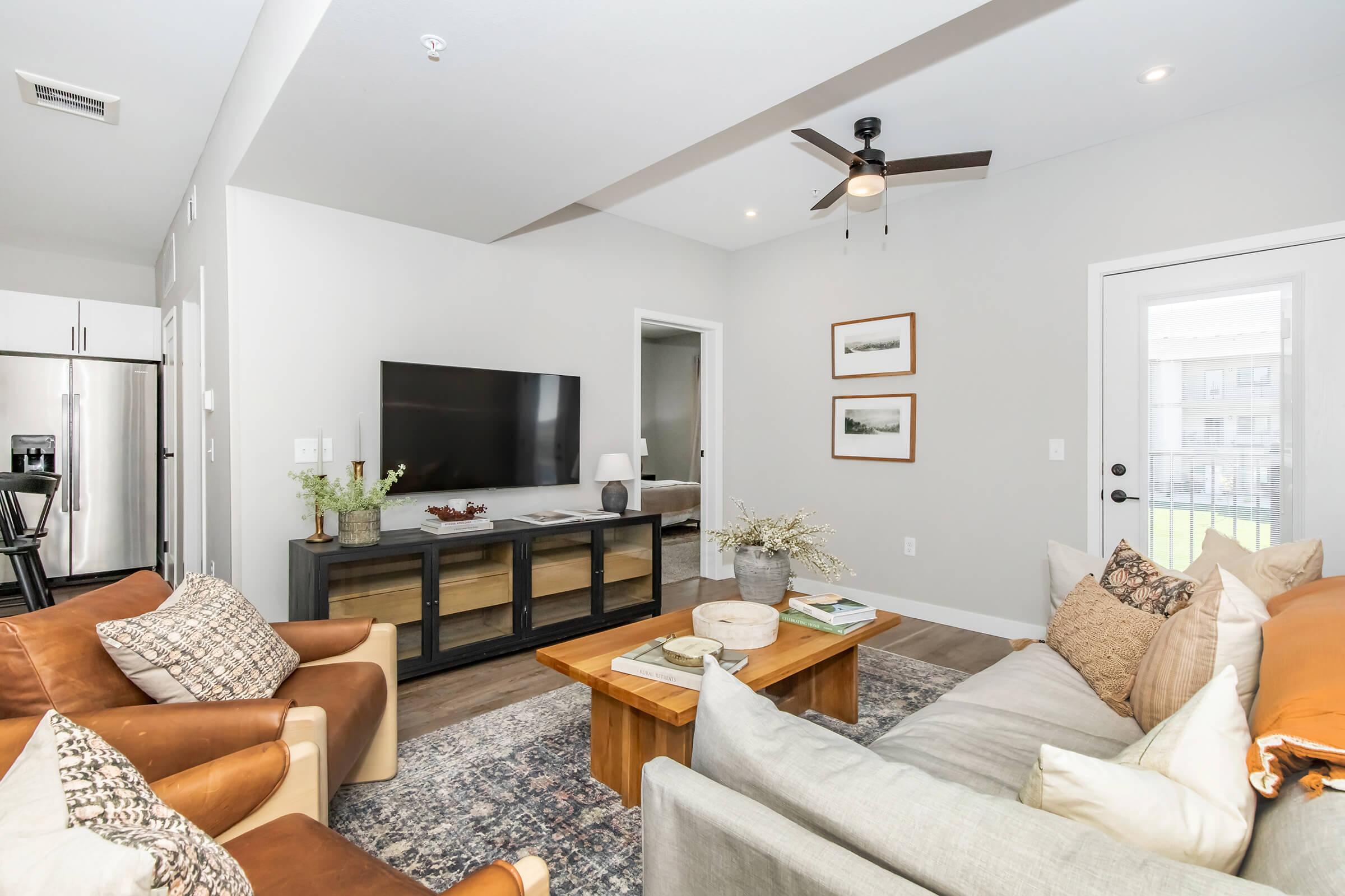 A modern living room with a light gray color scheme. It features a comfortable sofa with decorative pillows, an armchair, a wooden coffee table, and a TV unit. A ceiling fan hangs above, and the space is decorated with potted plants and wall art. Large windows provide natural light, creating a welcoming atmosphere.