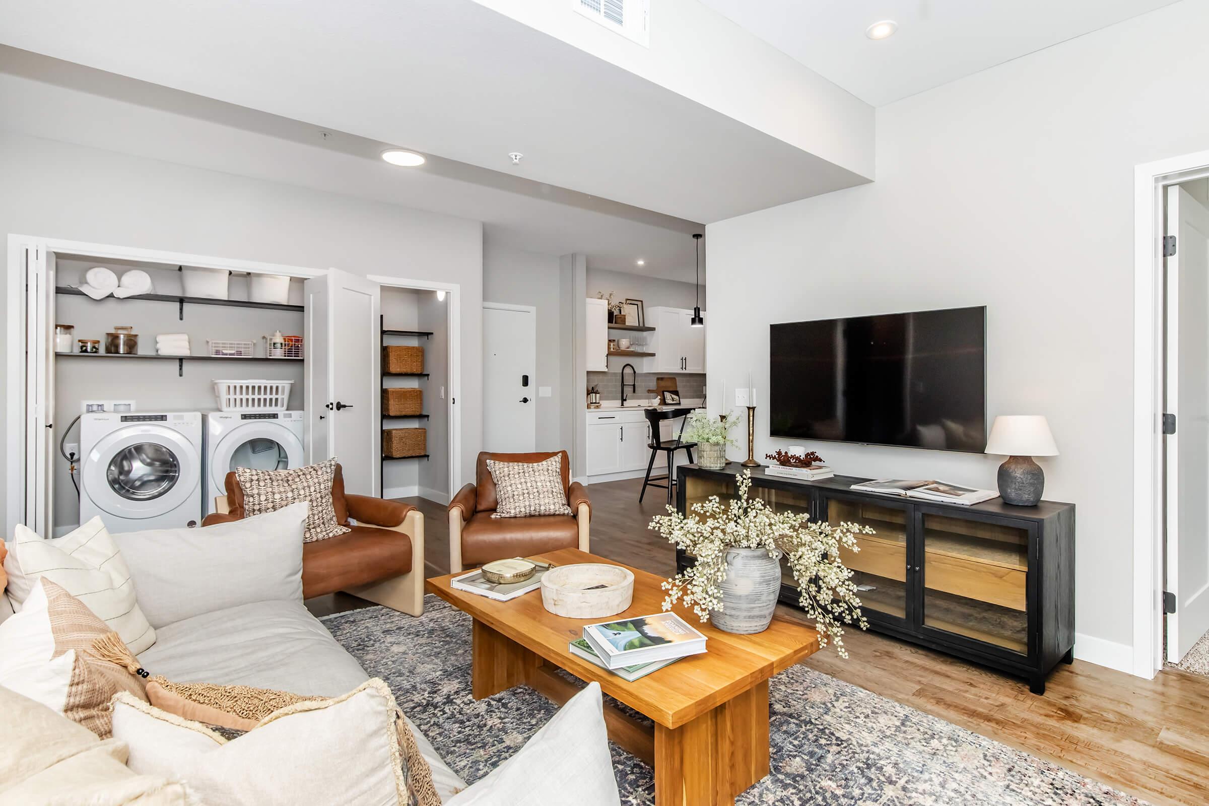 A modern living room featuring a cozy seating area with beige sofas and wooden chairs. A coffee table holds decorative items, while a TV is mounted on the wall. To the side, there are laundry appliances and shelves, enhancing the functional decor in a bright, open space with neutral tones.