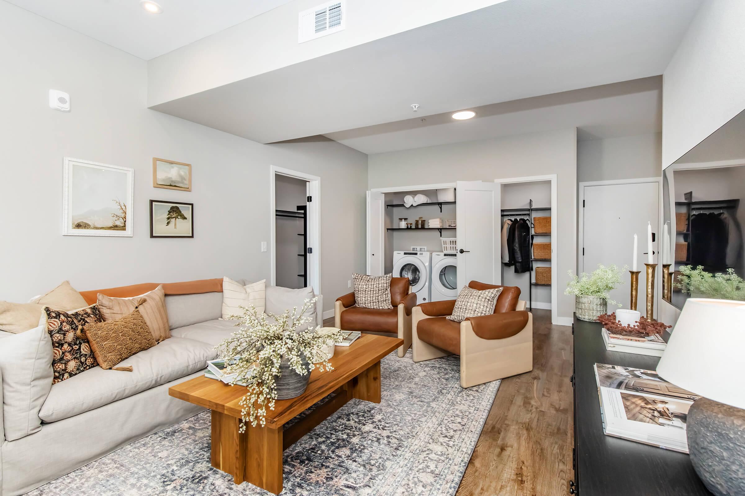 A stylish modern living room featuring a light-colored sectional sofa with decorative pillows, two brown accent chairs, a wooden coffee table with flowers, and a rug. In the background, there is a laundry area with a washer and dryer, and a wardrobe with clothes. The walls are adorned with framed art.