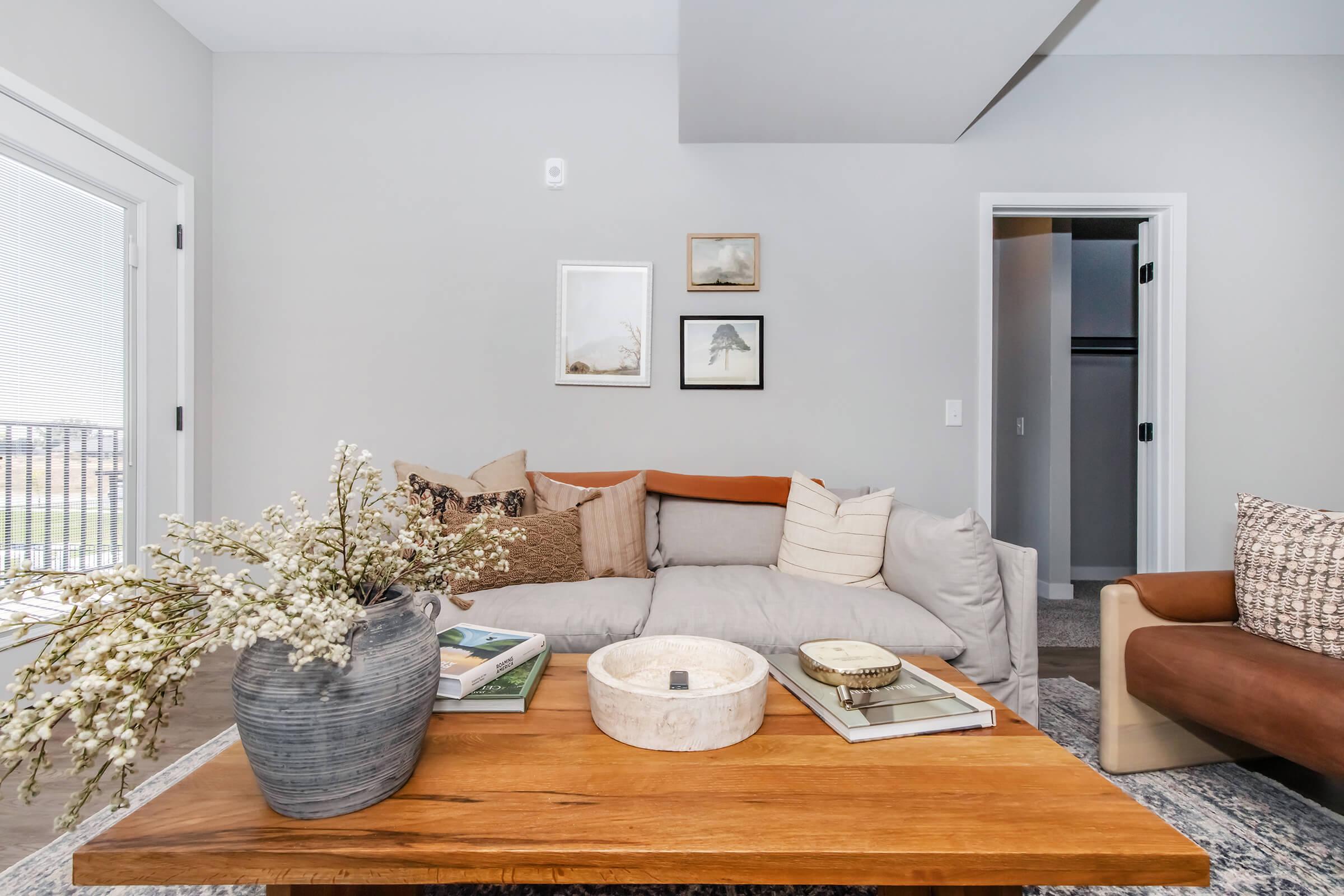 A cozy living room featuring a comfortable, light-colored sofa with decorative pillows. A wooden coffee table holds a plant vase and books. Light gray walls create a serene ambiance, with framed art on the walls and a doorway leading to another room.