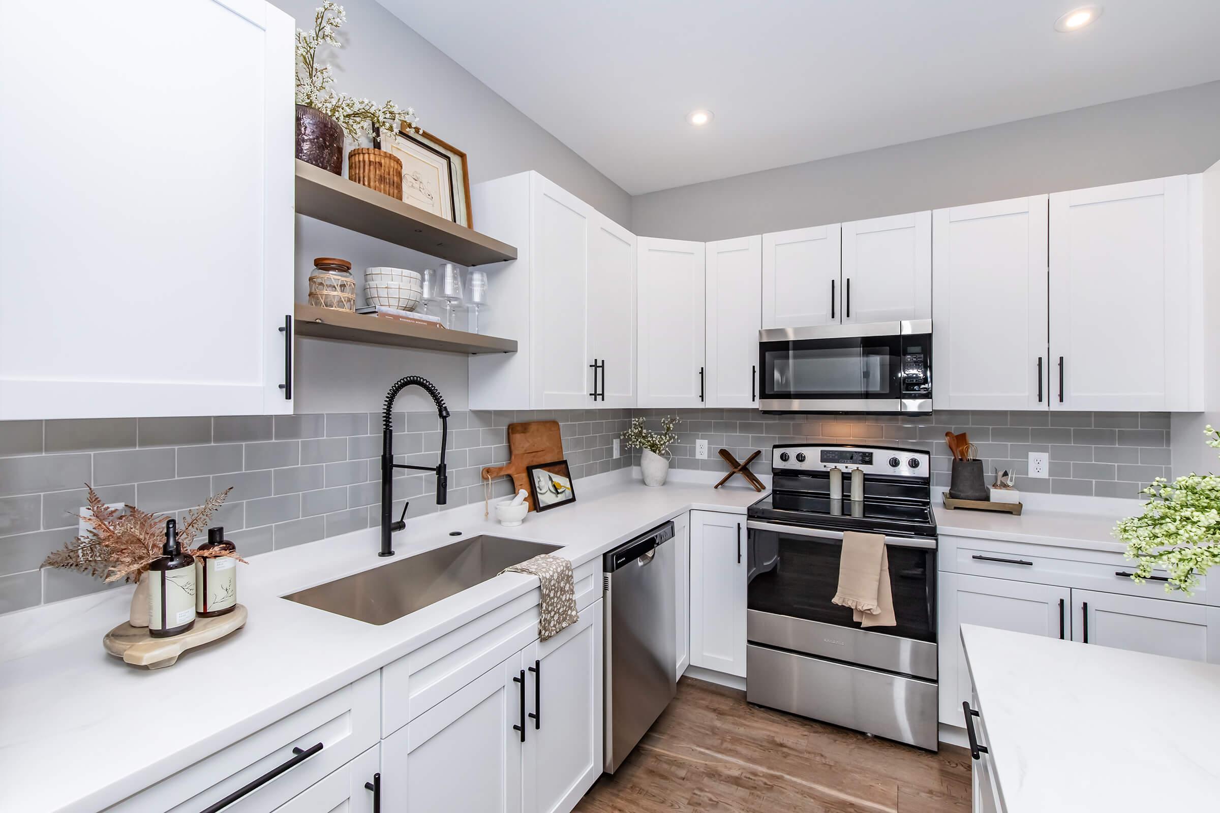 Modern kitchen featuring white cabinetry, stainless steel appliances, a large sink, and a sleek countertop. Décor includes wooden cutting boards, plants, and a few decorative items on open shelves, with muted gray tile backsplash and warm wood flooring.