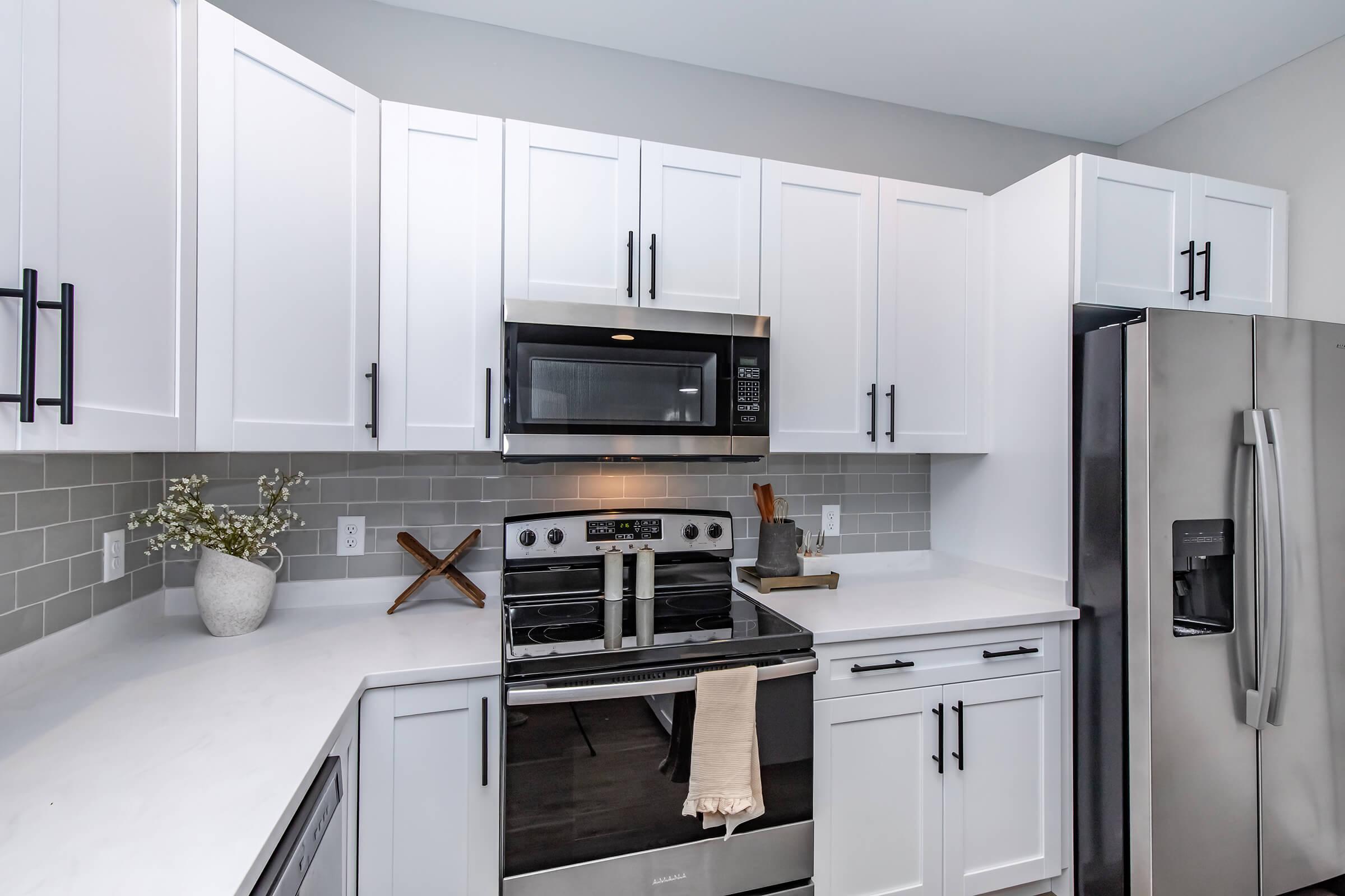 Modern kitchen with white cabinetry and stainless steel appliances. The countertop is light-colored, and a decorative vase with flowers is placed near the stove. A wooden cross is positioned on the countertop, and the refrigerator is sleek and silver. The overall design is clean and contemporary.