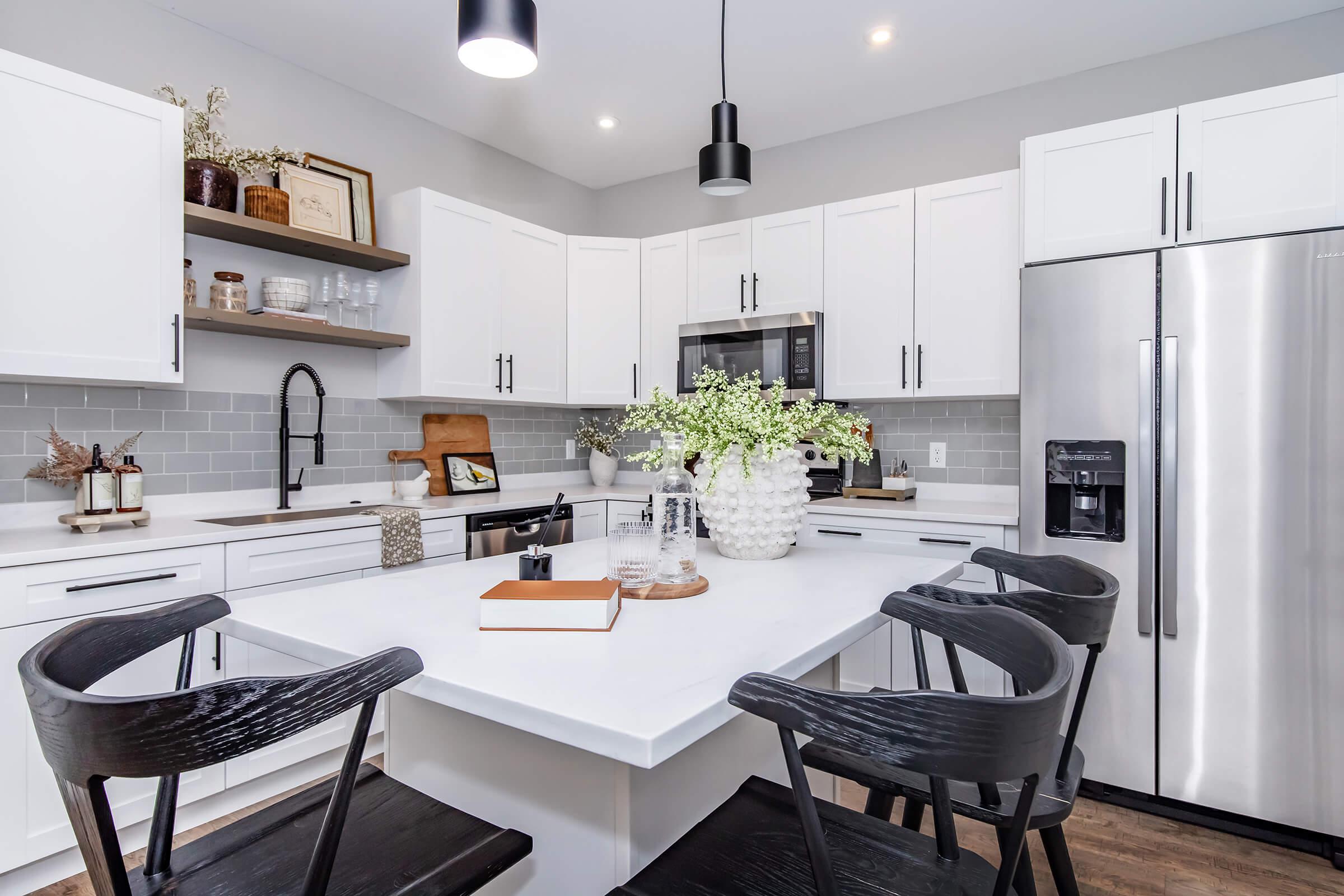 Modern kitchen featuring white cabinetry, a large island with decorative vase and books, black wooden chairs, stainless steel appliances, and a gray tiled backsplash. Natural light brightens the space.