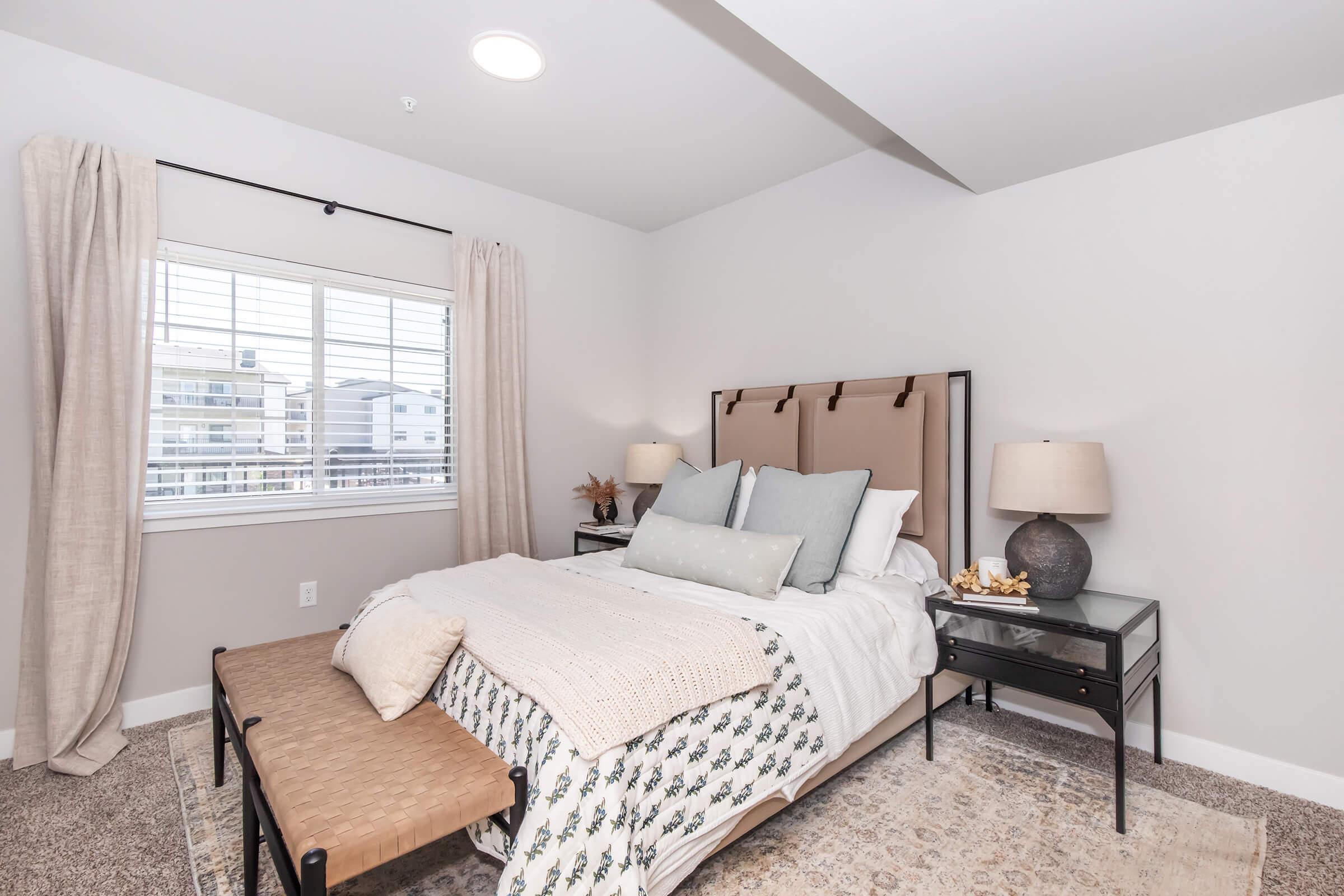 A modern bedroom featuring a beige upholstered bed with decorative pillows, a knitted throw blanket, and a stylish bench at the foot of the bed. There are two bedside tables with lamps, and large windows with light curtains, illuminating the neutral-colored walls and carpeted floor.