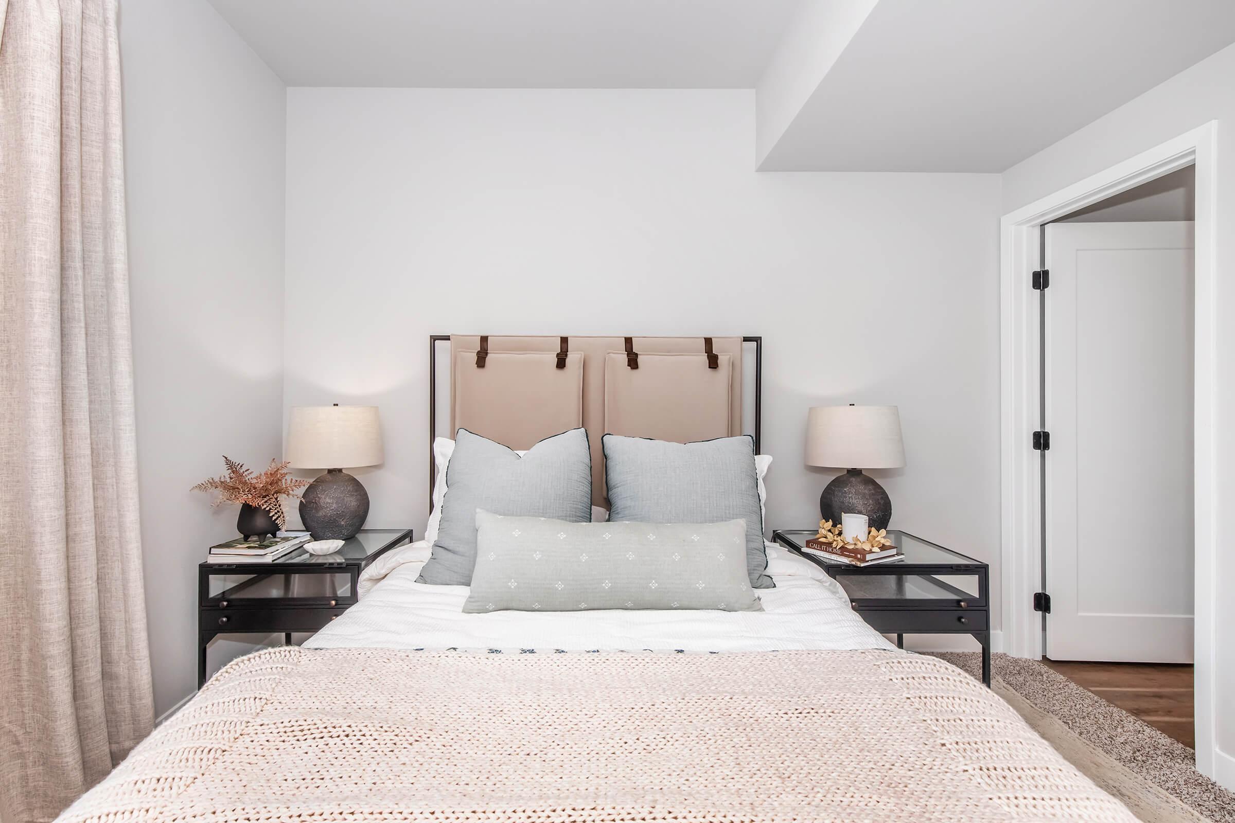 A cozy bedroom featuring a neutral color palette. The bed has a headboard with brown straps, adorned with light gray and white pillows. Side tables with dark lamps flank the bed, and a textured blanket lies across the foot. A door leads to another room, while light curtains frame the window.