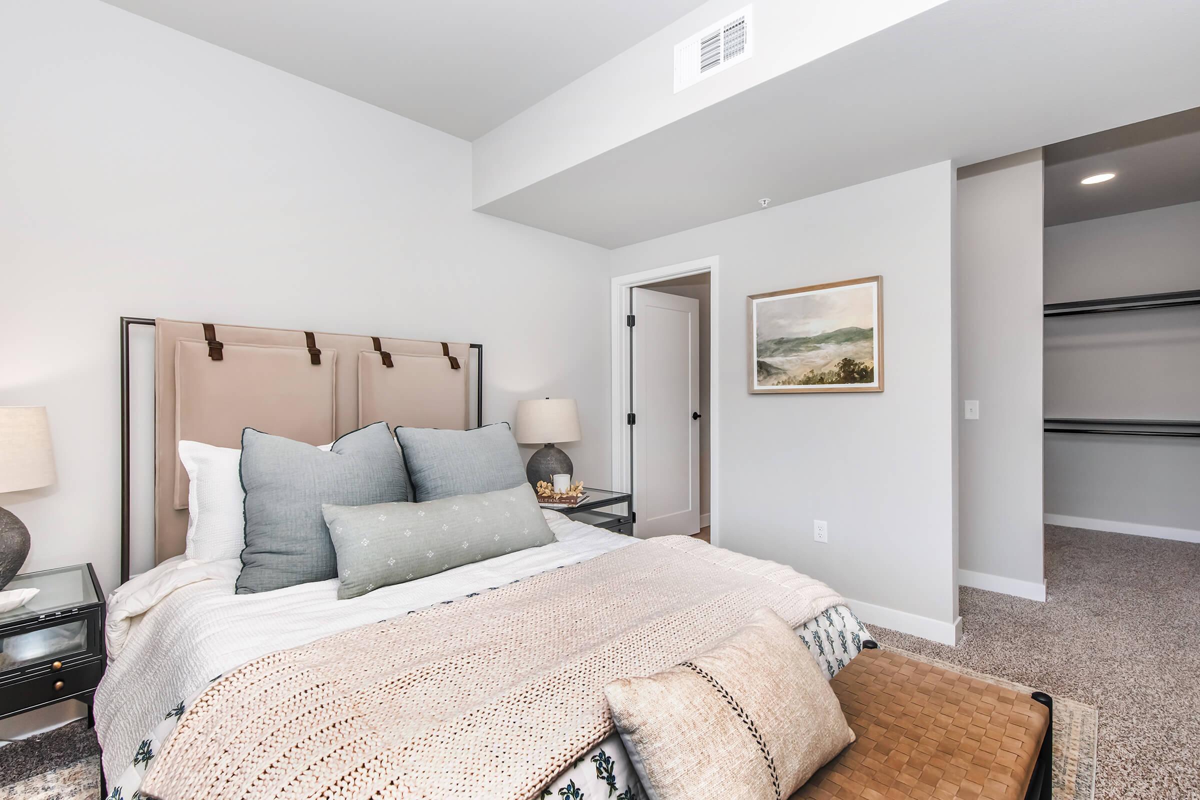 A modern bedroom featuring a neatly made bed with plush pillows and a cozy blanket. There are bedside tables with lamps, a framed landscape picture on the wall, and a spacious closet area. The room has soft, neutral colors and a clean, inviting aesthetic.