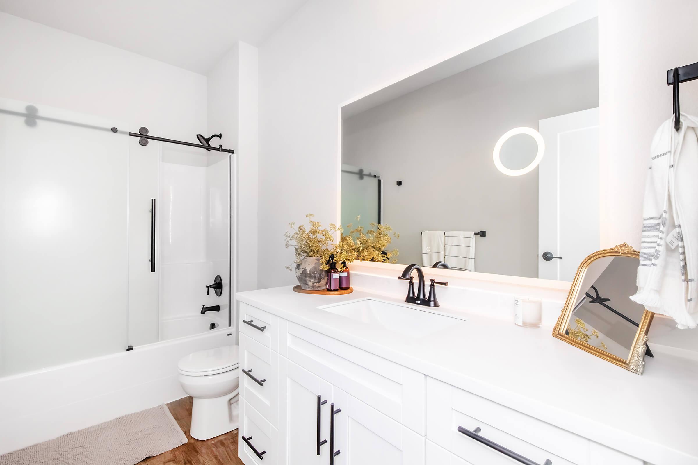 Modern bathroom featuring a sleek white vanity with black fixtures, a large mirror, and a cozy decor element. The space includes a bathtub with a glass shower door, a toilet, and decorative items like a soap dispenser and plants, creating a clean and inviting atmosphere.