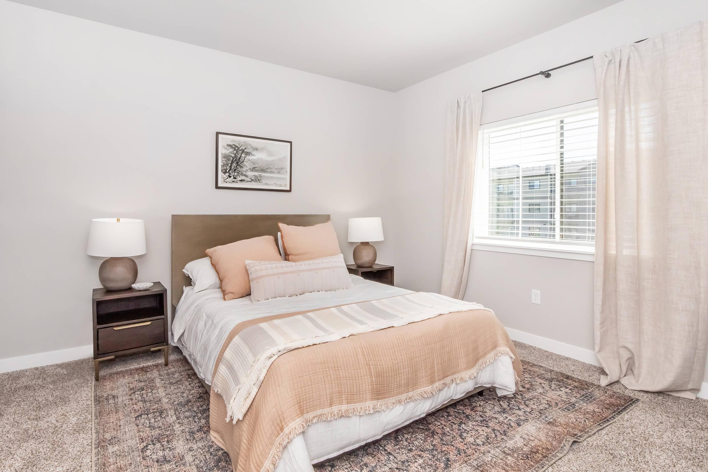 Cozy bedroom featuring a soft color palette with a neatly made bed adorned with light brown and cream bedding. There are two bedside tables with lamps, a window with sheer curtains allowing natural light, and a piece of framed wall art above the bed, complemented by a textured area rug.