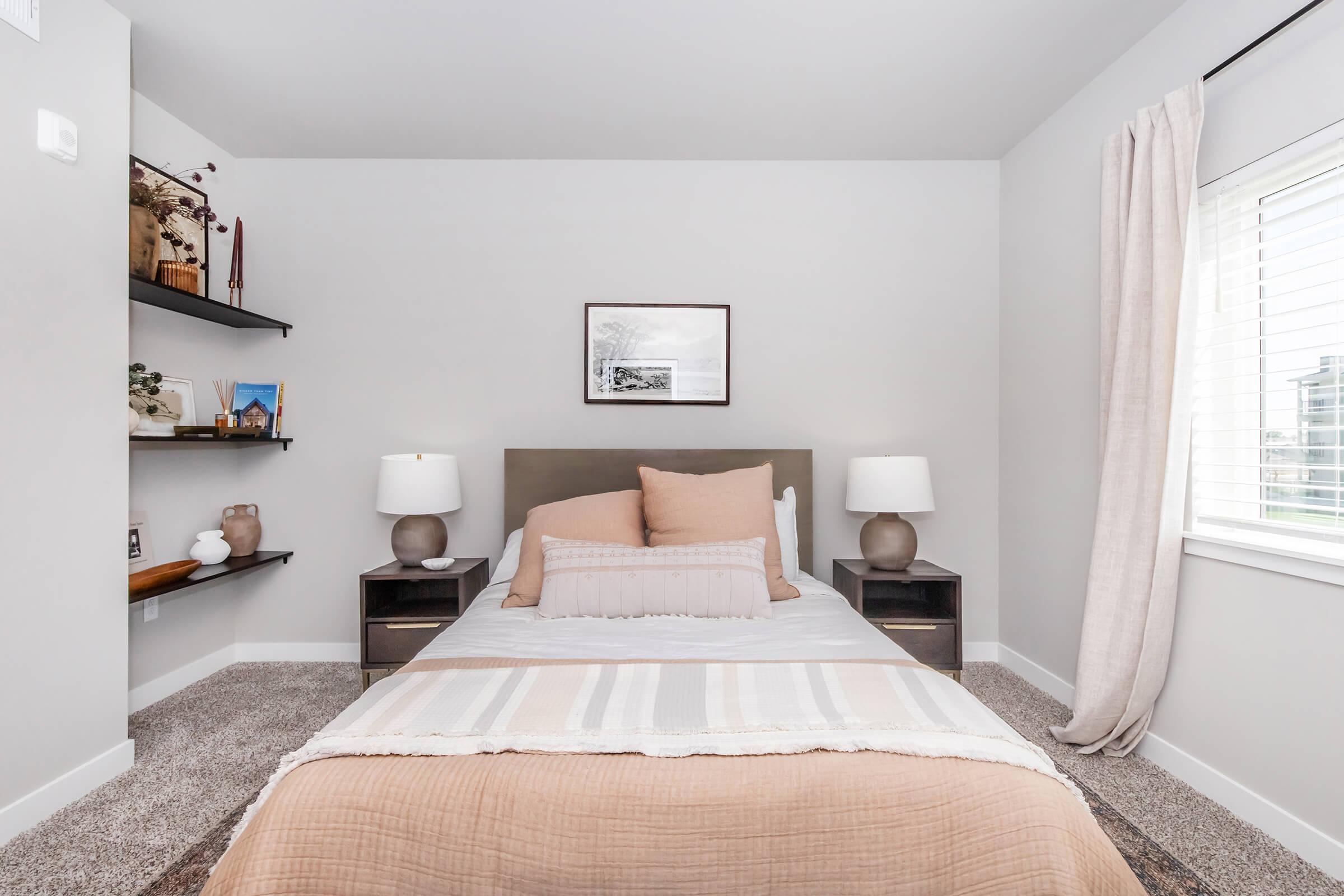 A cozy bedroom featuring a comfortable bed with beige and pink pillows, two nightstands with lamps, and a simple wall frame above the bed. There are shelves on the wall displaying decorative items, and the room has soft lighting and neutral-colored walls, creating a calm atmosphere.