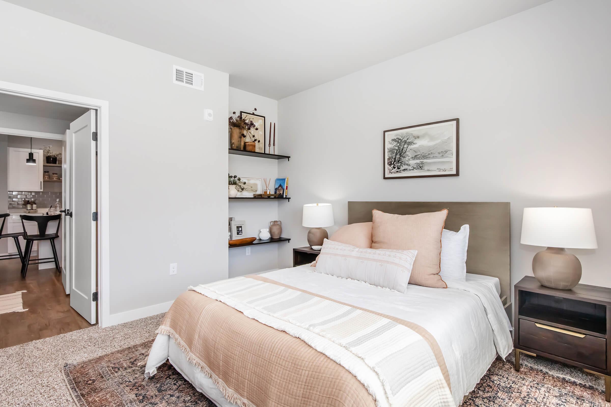 A cozy bedroom featuring a queen-sized bed with neutral bedding, two bedside lamps, and a simple decorative painting on the wall. A shelf displays decorative items, while a doorway leads to an adjoining room. The room has light-colored walls and a soft area rug on the wooden floor.