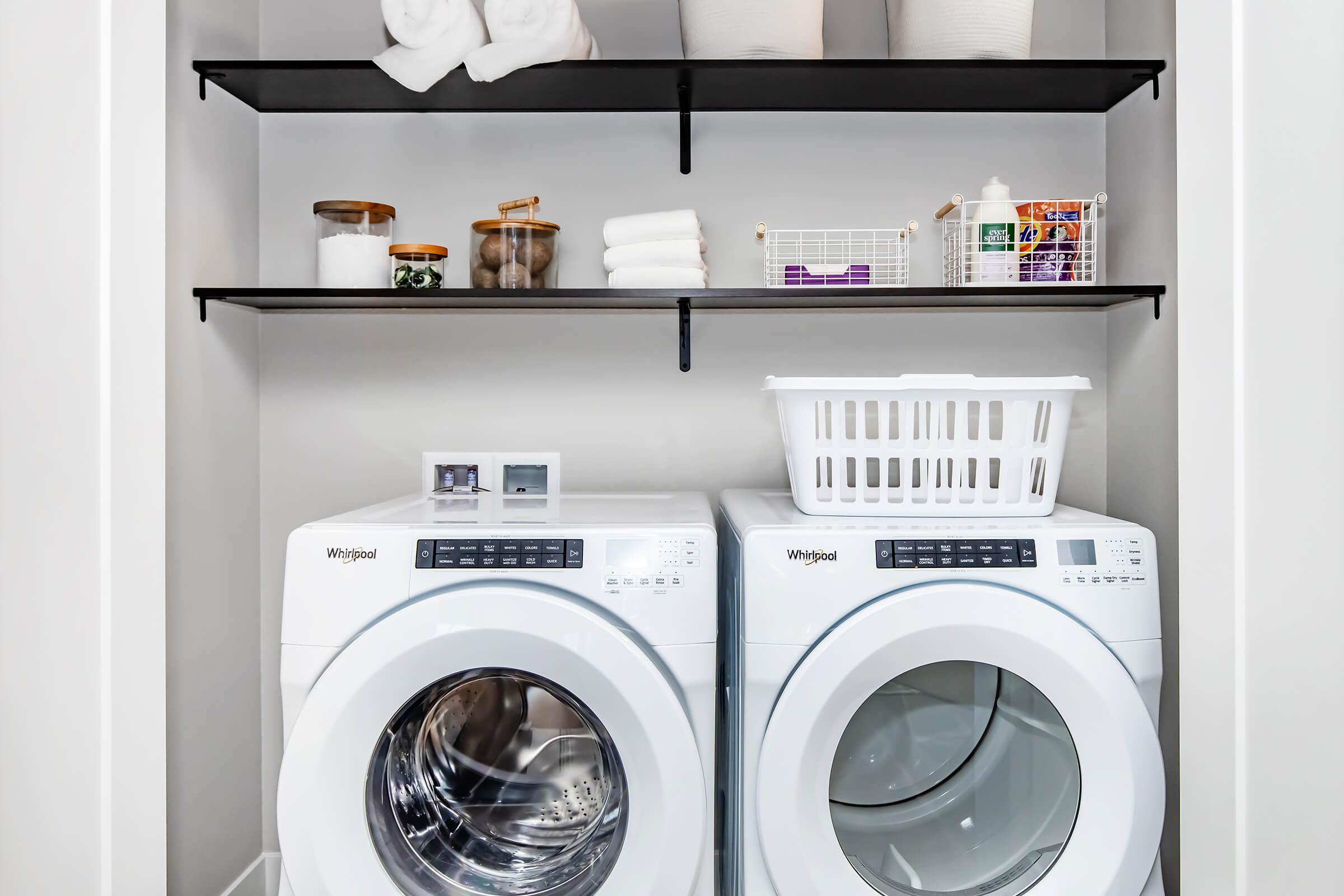 A modern laundry room featuring two white Whirlpool washing machines side by side. Above them, two black shelves display neatly arranged towels, storage jars, and cleaning supplies. A white laundry basket is positioned in front of the machines, creating a clean and organized space.